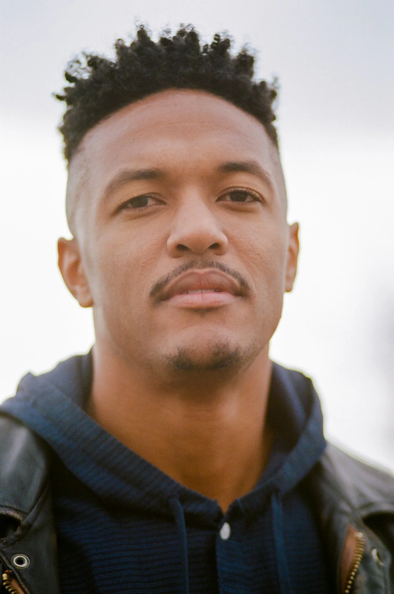 Close-up portrait of a man with short curly hair, light mustache, wearing a navy hoodie and leather jacket, outdoors with a cloudy sky in the background.