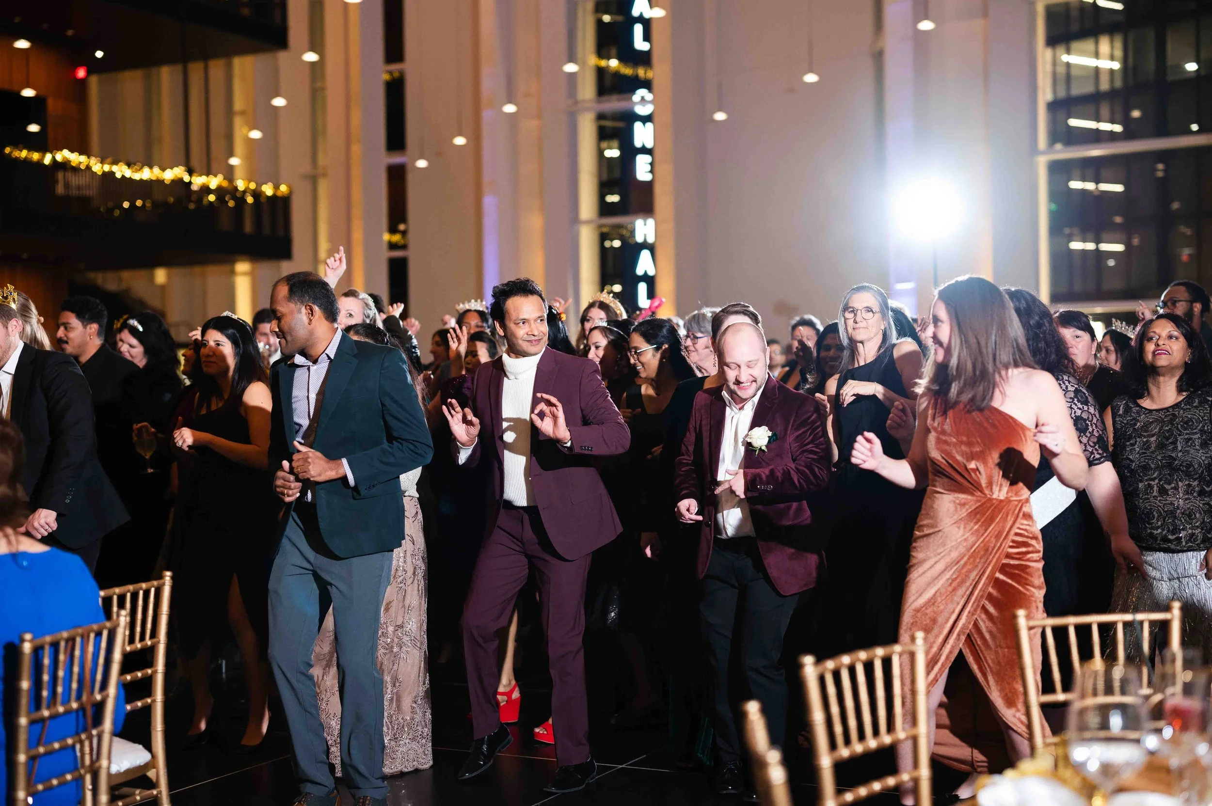 Group of people dancing at an indoor event, dressed in formal attire, with festive lighting and decorations.