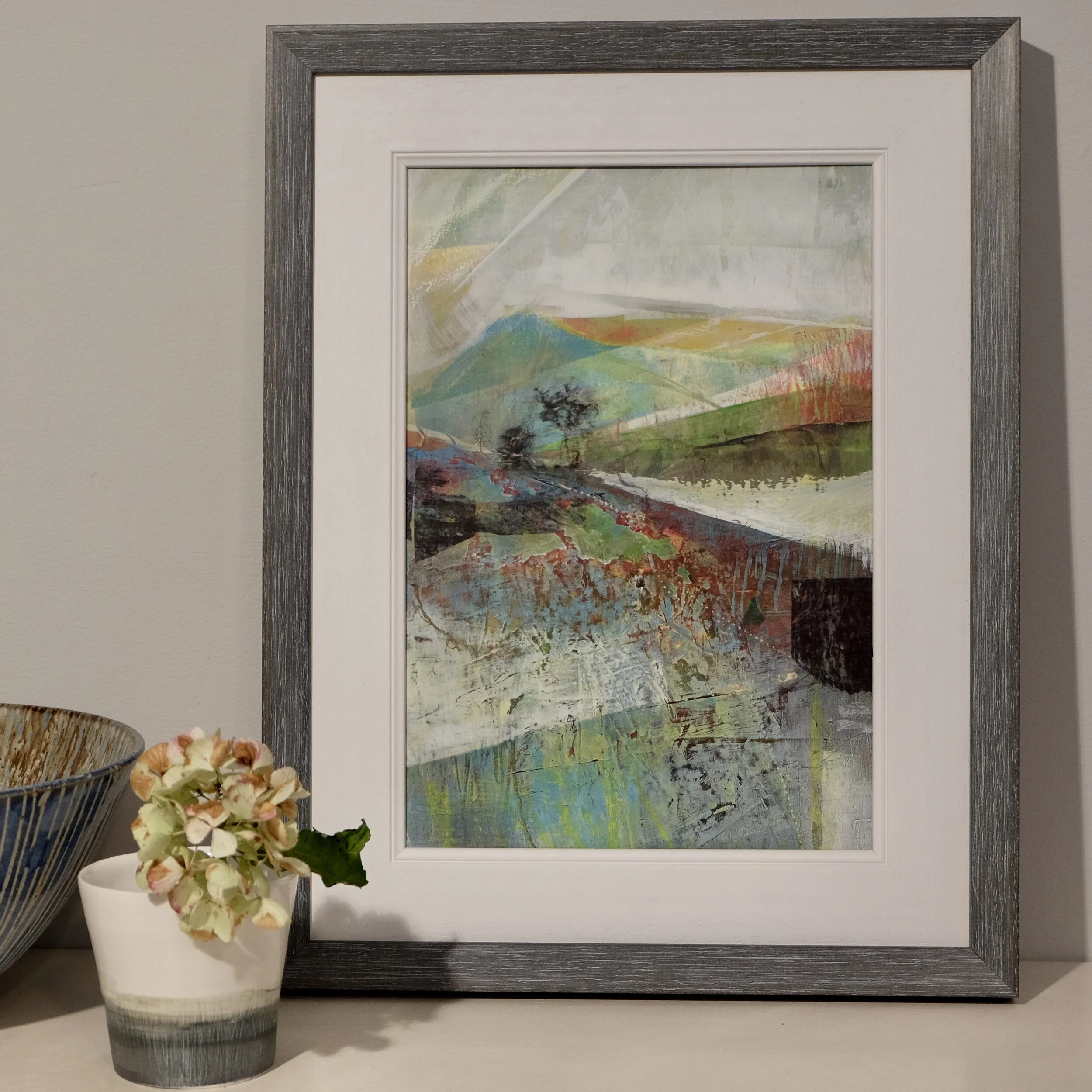 Framed acrylic landscape painting with hills and trees by Joanne Olney displayed with flower filled vessel