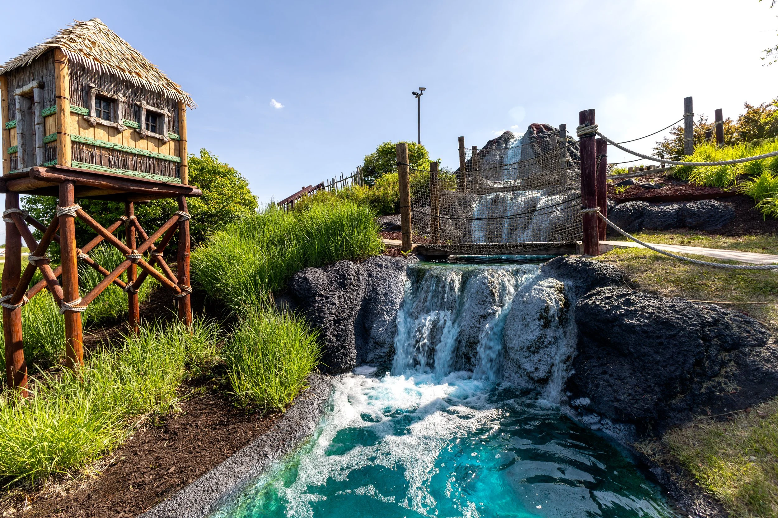 A small waterfall flowing down rocks in a natural setting with a wooden fence and a thatched-roof hut on stilts in the background, during daytime with clear blue sky.