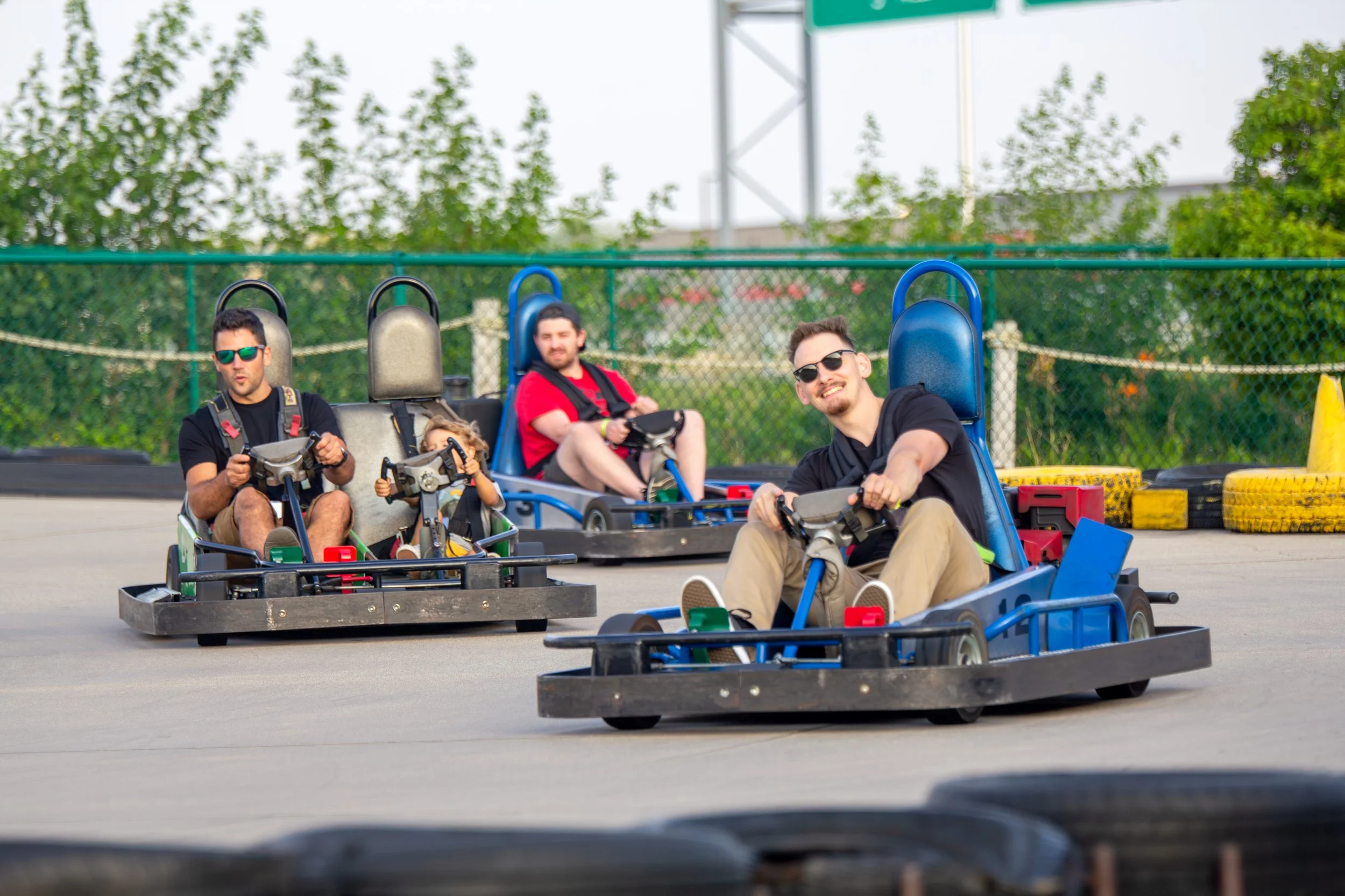 Four people riding go-karts outdoors on a track with a background of trees and a fence, wearing sunglasses and casual clothing.