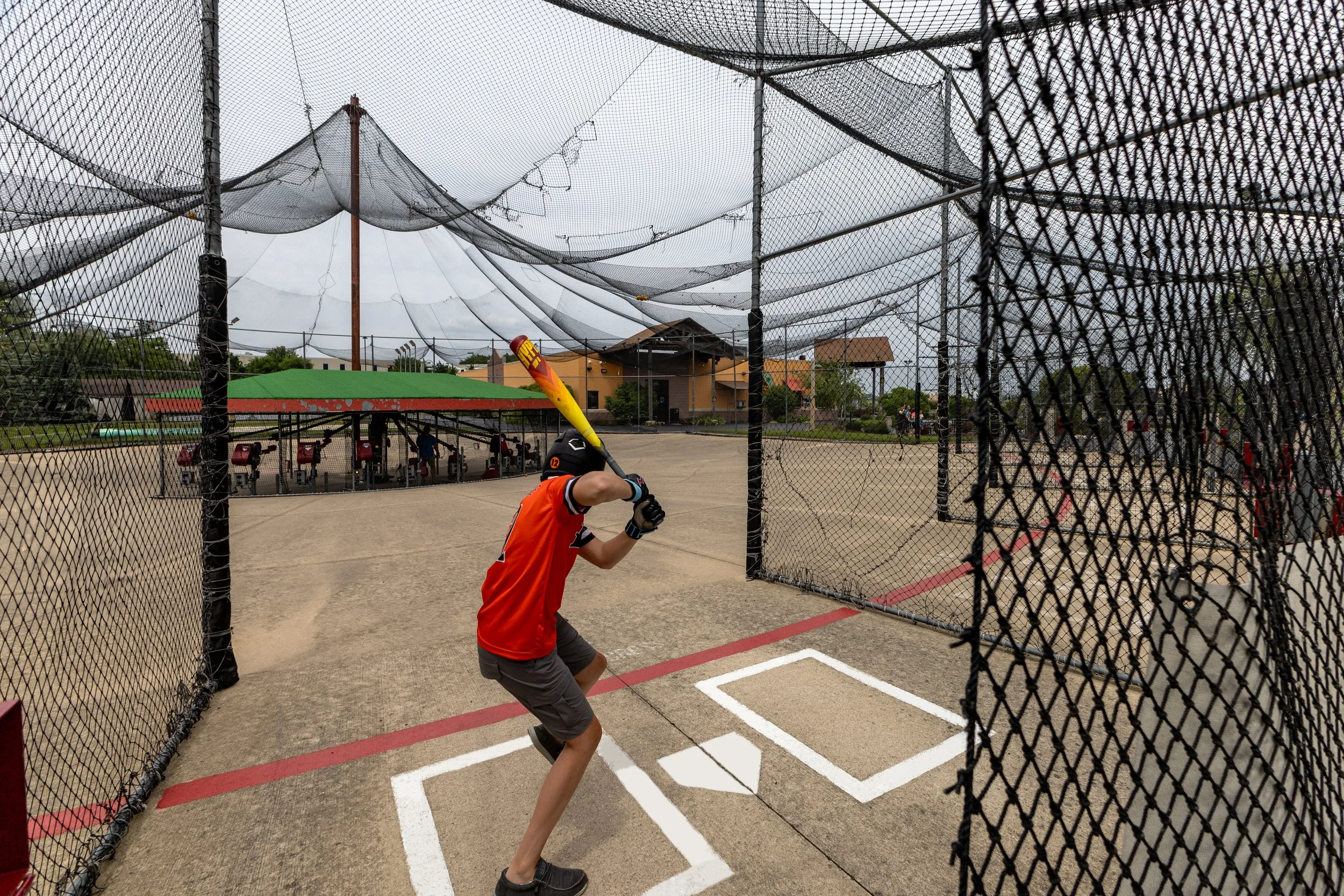 Child in an orange shirt playing baseball in a batting cage with nets overhead and surrounding the field.