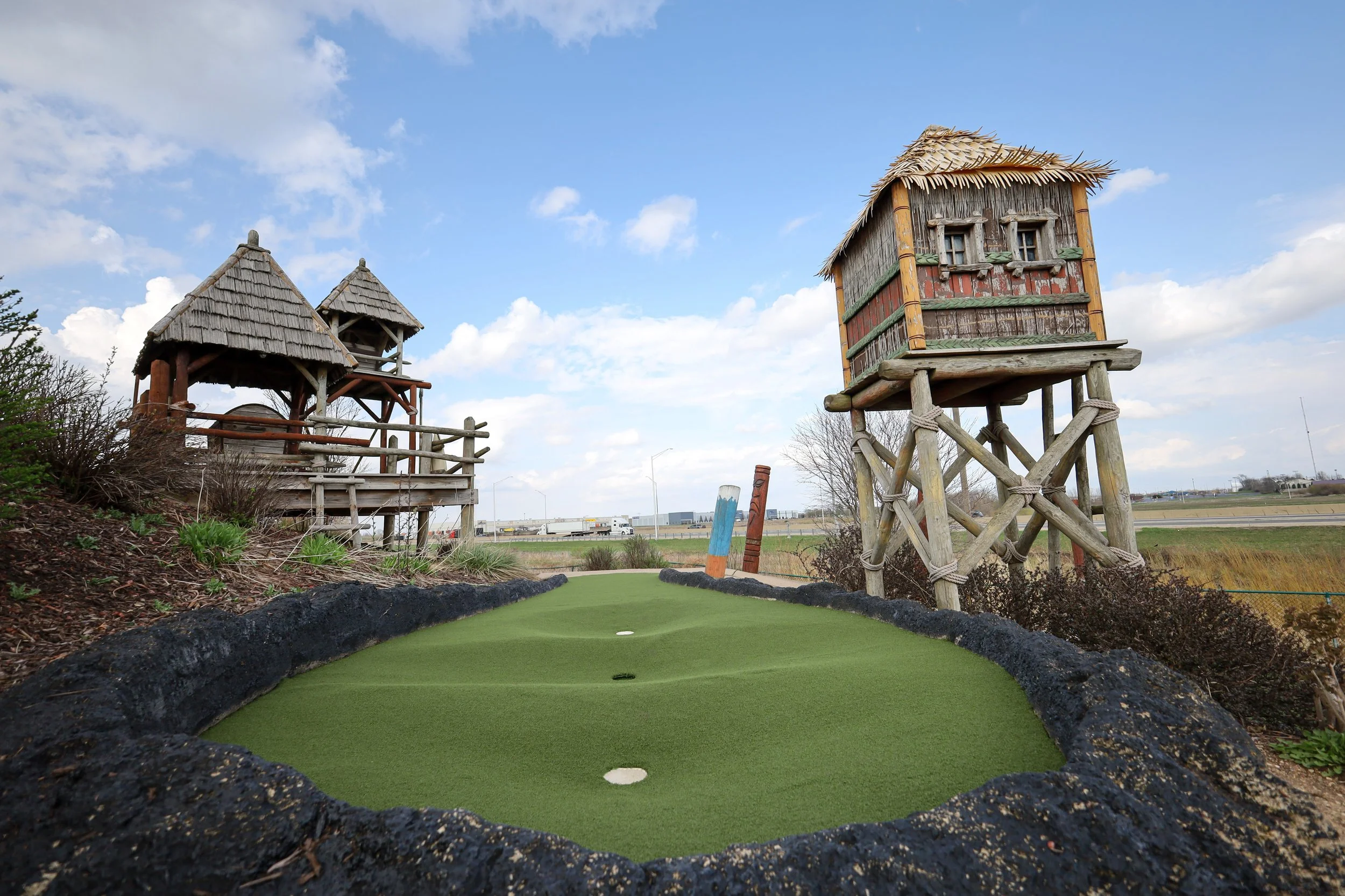 Miniature golf course with a putting green, rocks along the edges, and tiki-style huts and towers in the background under a partly cloudy sky.