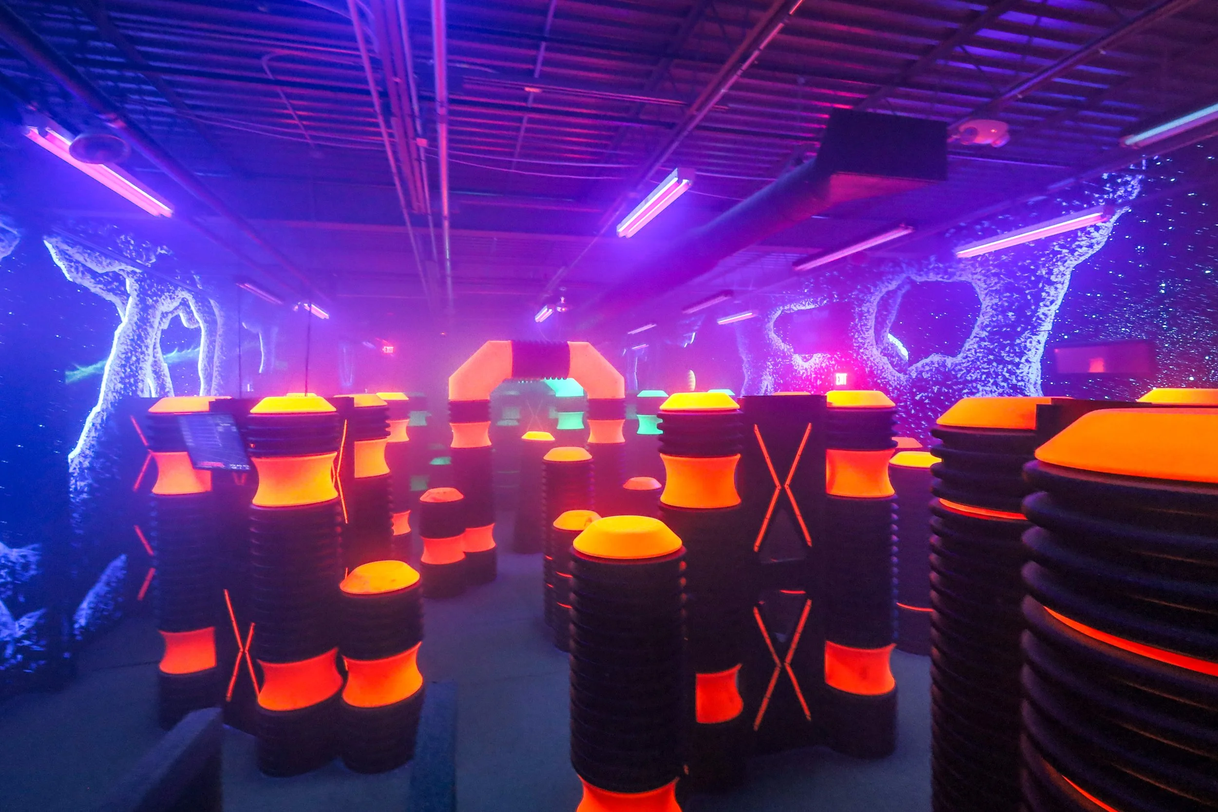 Colorful arcade or entertainment space with neon lighting, abstract backdrop, and stacked orange and black stools.