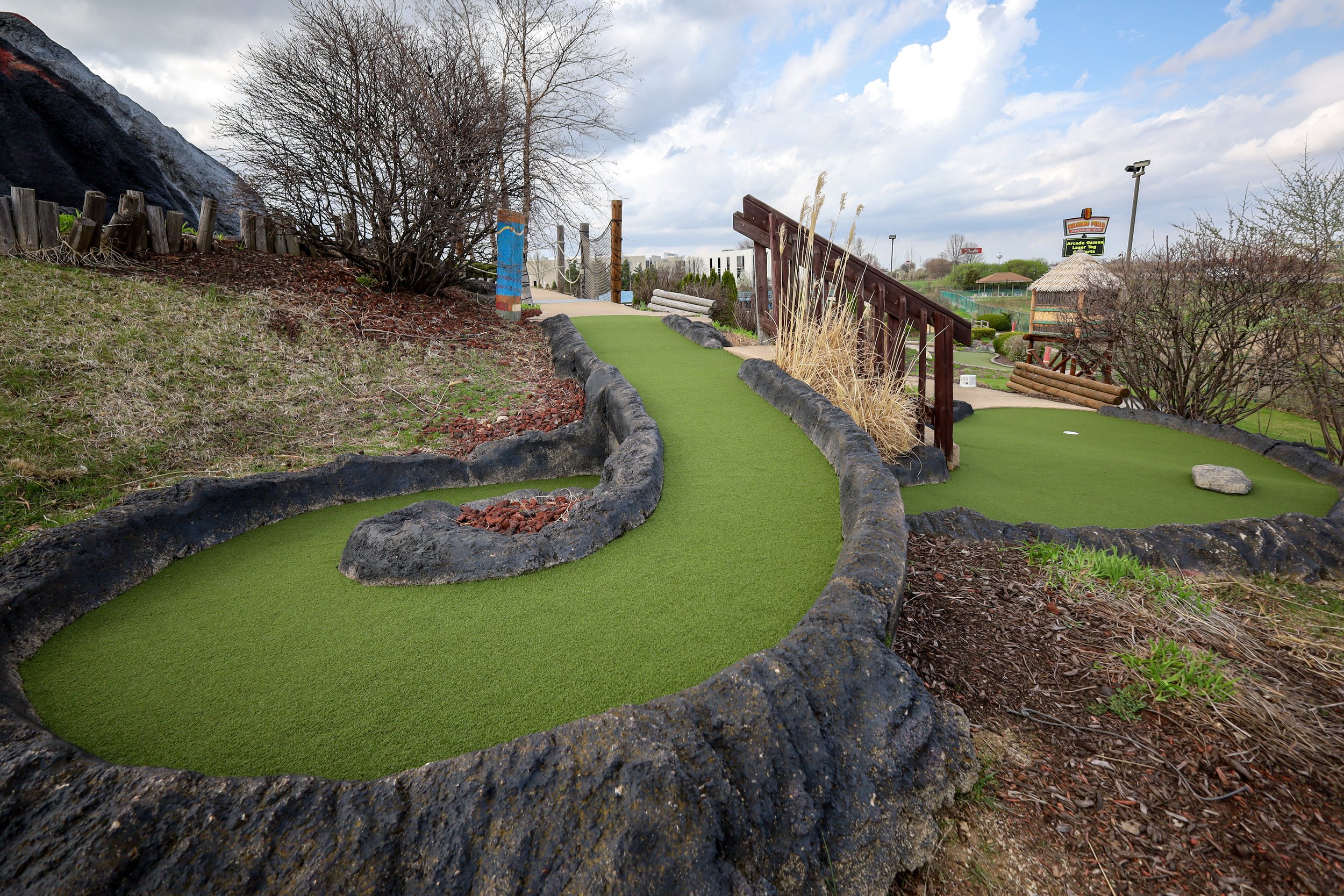 Miniature golf course with artificial turf and black rubber borders, surrounded by landscaping, trees, and a wooden bridge, with a cloudy sky overhead.