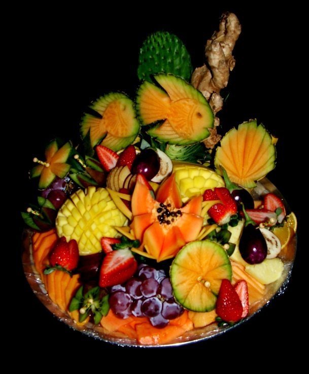 Colorful fruit platter with sliced strawberries, grapes, pineapple, melon, papaya, kiwi, and a carved fruit flower, garnished with ginger and decorative leafy accents, set against a black background.