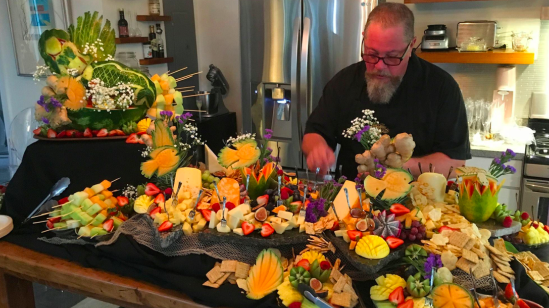 Man preparing a large elaborate fruit and cheese display on a kitchen counter