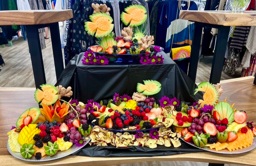 Assorted fresh fruit platter display featuring pineapple, strawberries, blackberries, blueberries, grapes, and melon slices, arranged on three large trays with decorative fruit art.