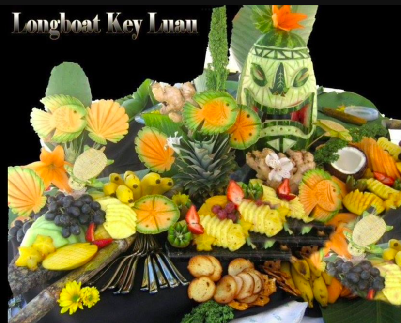 Tropical fruit display including pineapple, bananas, grapes, strawberries, and assorted sliced fruits, decorated with paper umbrellas, flowers, and a tiki mask.