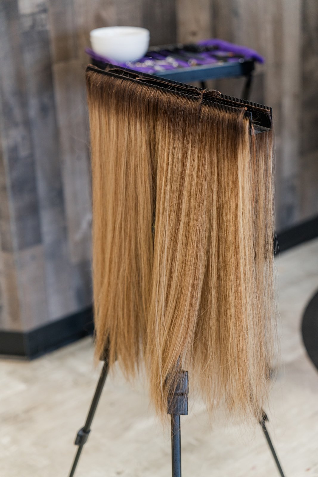 Blonde hair extensions hanging on a stand, with a bowl and styling tools in the background.