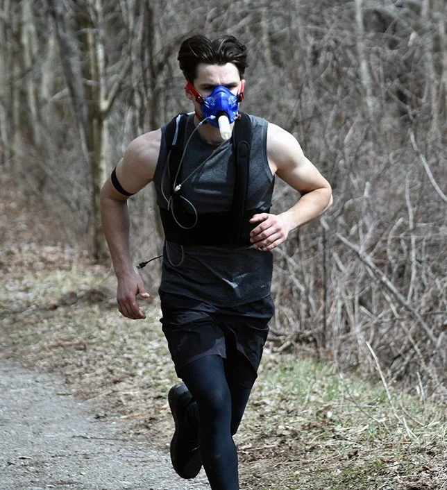 A man running outdoors on a trail, wearing a respirator mask, sleeveless shirt, running gear, and headphones, in a wooded area during daytime.
