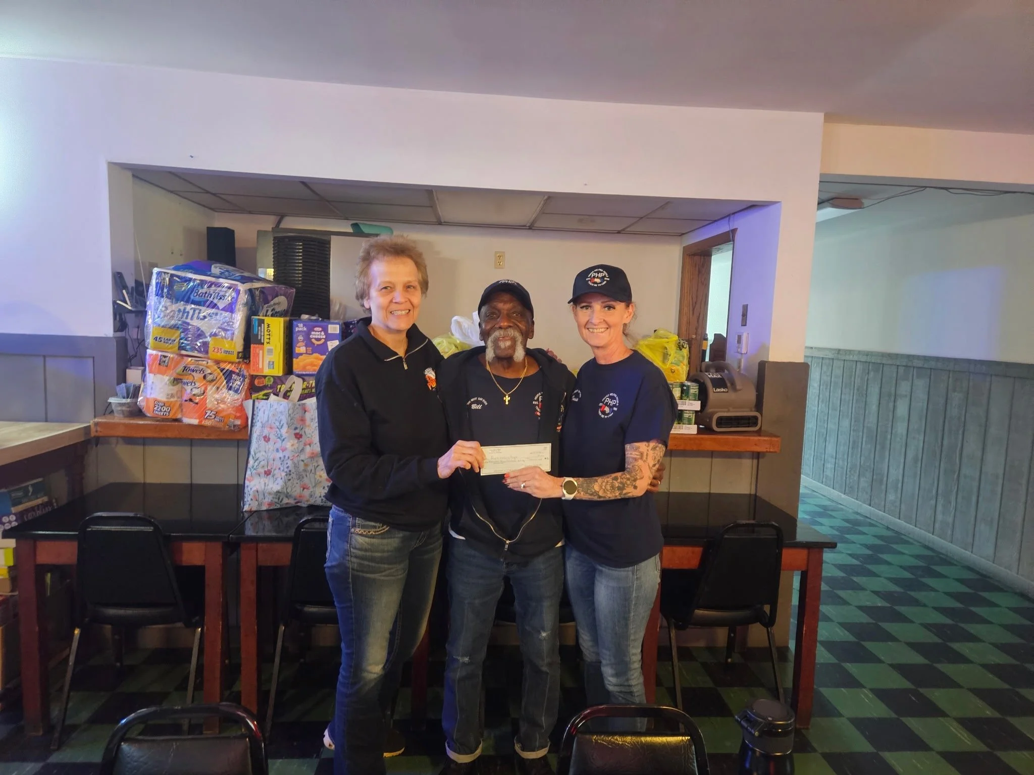 Three people in matching shirts hold a check during a community donation presentation, with donated paper products stacked behind them.