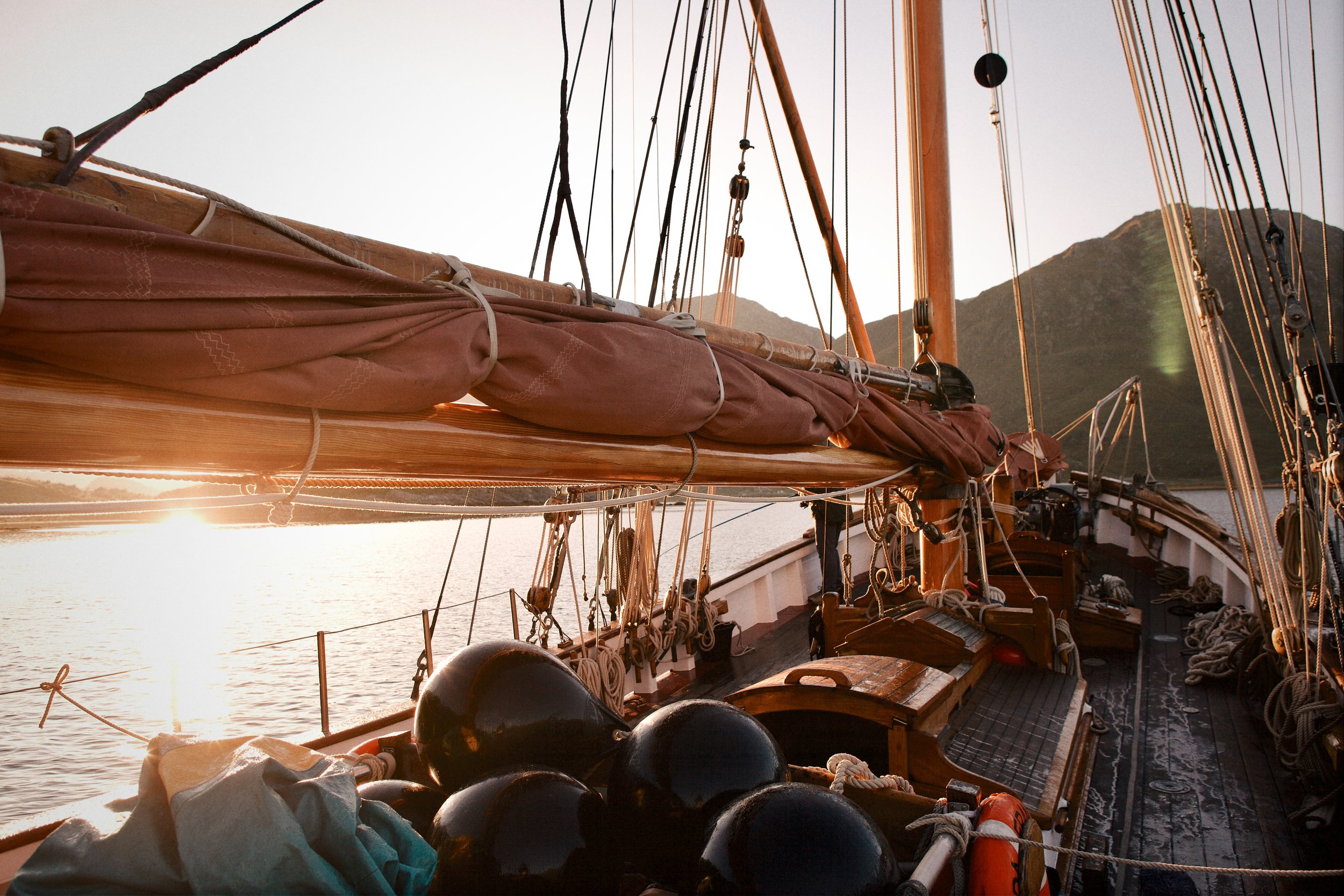 View of a sailboat's deck with ropes, black fenders, and wooden elements during sunset, with mountains in the background.