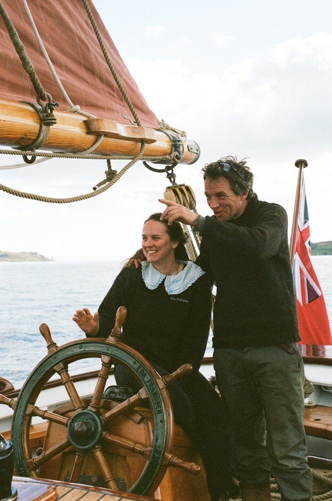 A smiling woman and man on a sailboat, with the man fixing the woman's hair. The boat's wooden steering wheel is in the foreground, and a Union Jack flag is visible on the right side of the boat. The ocean and sky are in the background.