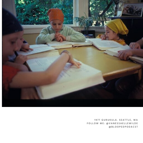 Four children wearing colorful head coverings sitting at a table reading books in a classroom with large windows and plants in the background.