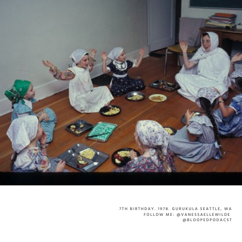 Children dressed in traditional clothing sitting on the floor in a circle around a woman, who appears to be telling a story or leading an activity, with food on trays in front of them.