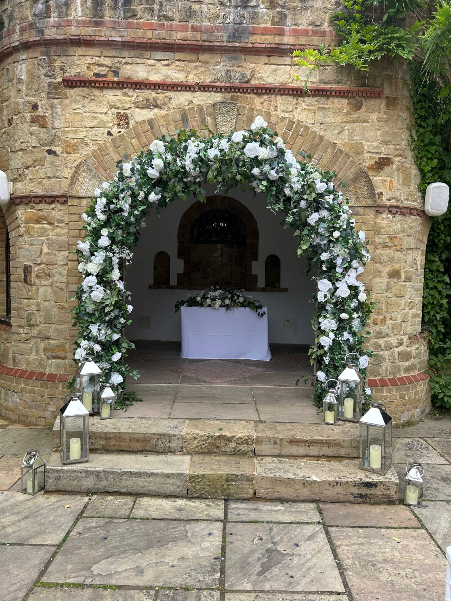 Luxury Floral Arch decorated with white flowers and greenery, available to hire for your wedding or event, set in front of a stone archway with lanterns on steps.