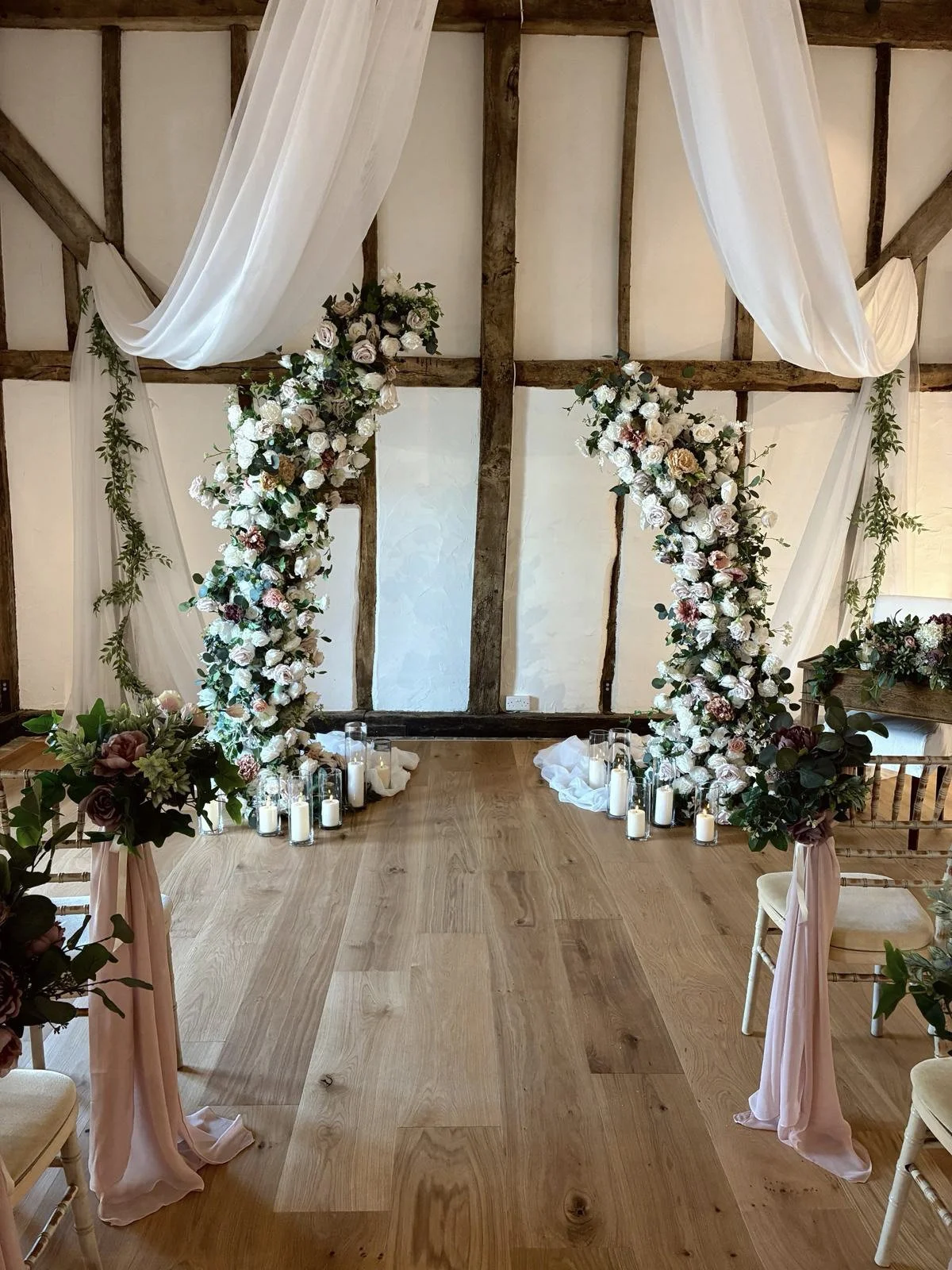 Asymetric/Broken Arch nude pink and white with floral and greenery design, surrounded by candles and draped fabric, available to hire for your wedding or event.