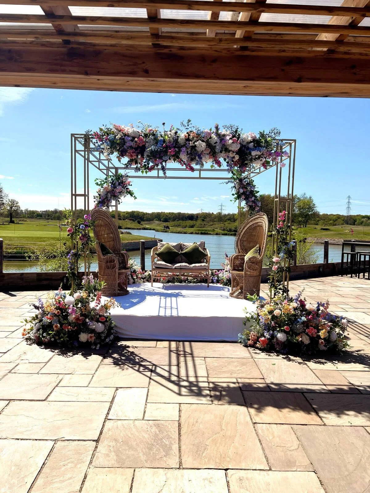 Mandap - Gold Metal decorated with pastel and purple flowers, wicker seating on white platform, available to hire for your wedding or event.