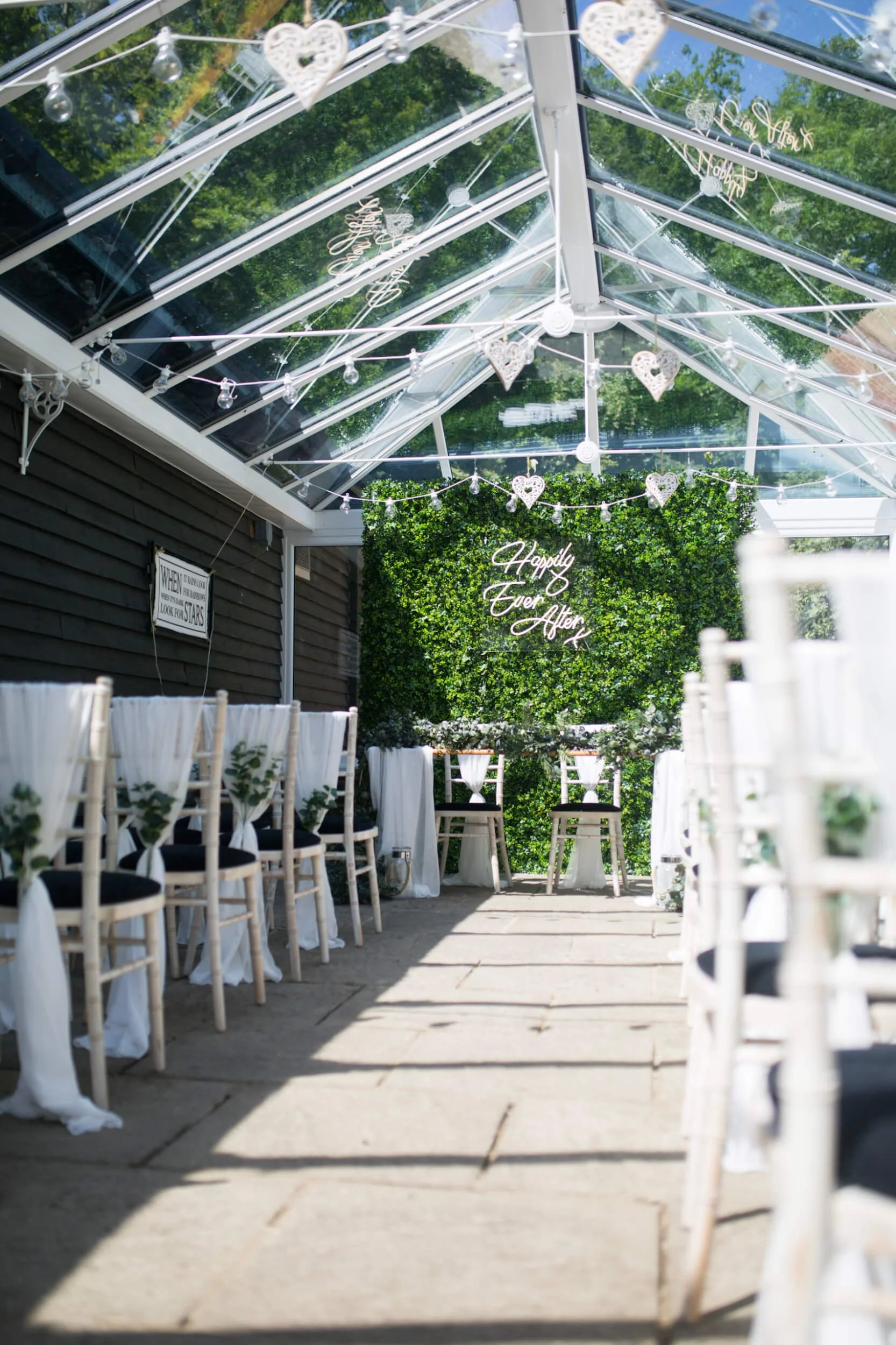 Foliage Wall with neon 'Happily Ever After' sign, available to hire for your wedding or event, set in a decorated aisle under a glass roof with hanging hearts and lights.