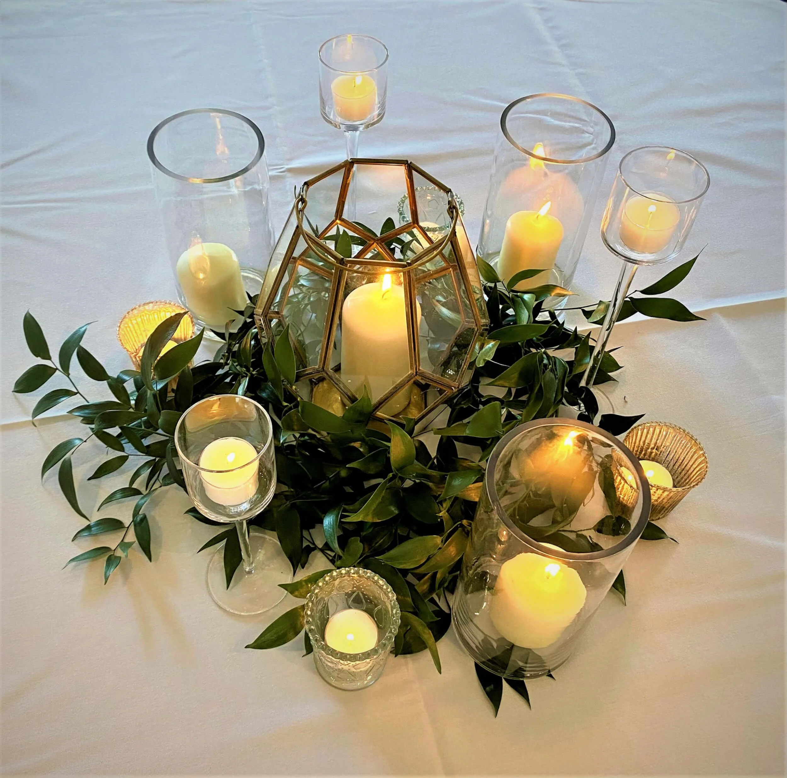 Foliage Mohani and glass centrepieces - Various Sizes with lit white candles and green foliage, available to hire for your wedding or event.