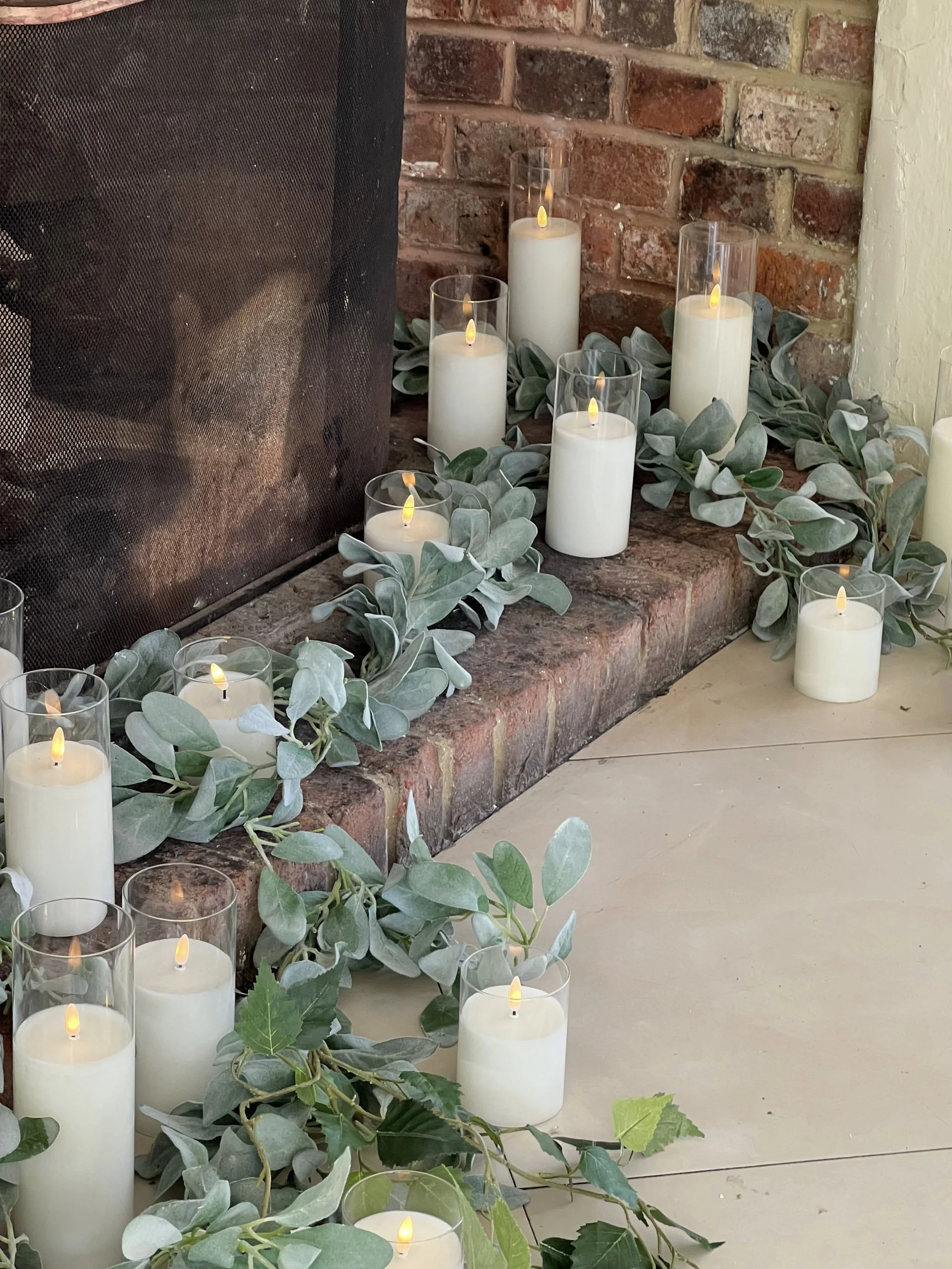 Clear Cylinder LED White Candle - Large 23cm high x 7 cm diam arranged on a brick hearth with greenery, available to hire for your wedding or event.