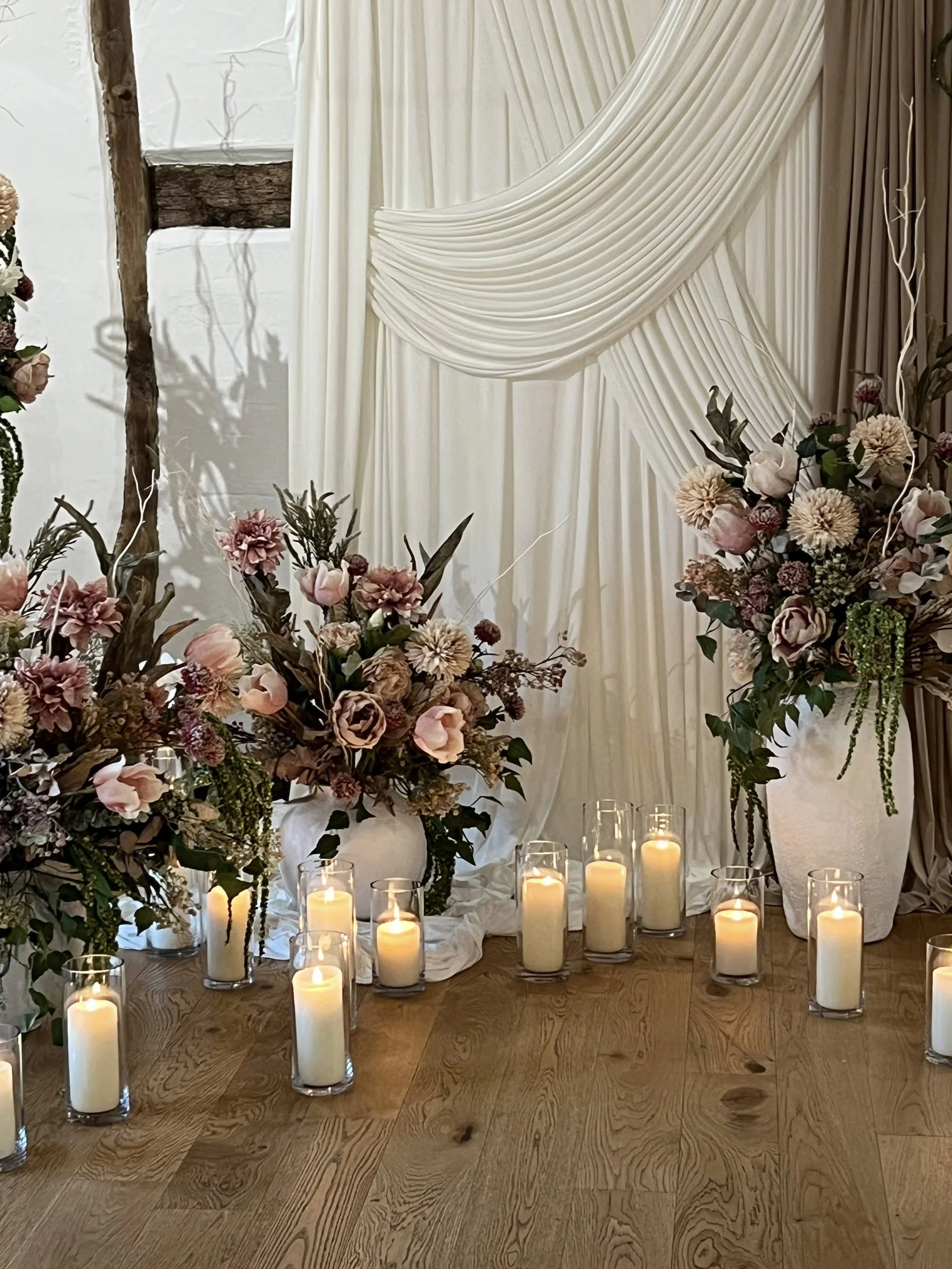 Cylinder Vase - 30cm with lit white pillar candles inside, available to hire for your wedding or event, arranged on wooden floor with floral decor and draped fabric backdrop.