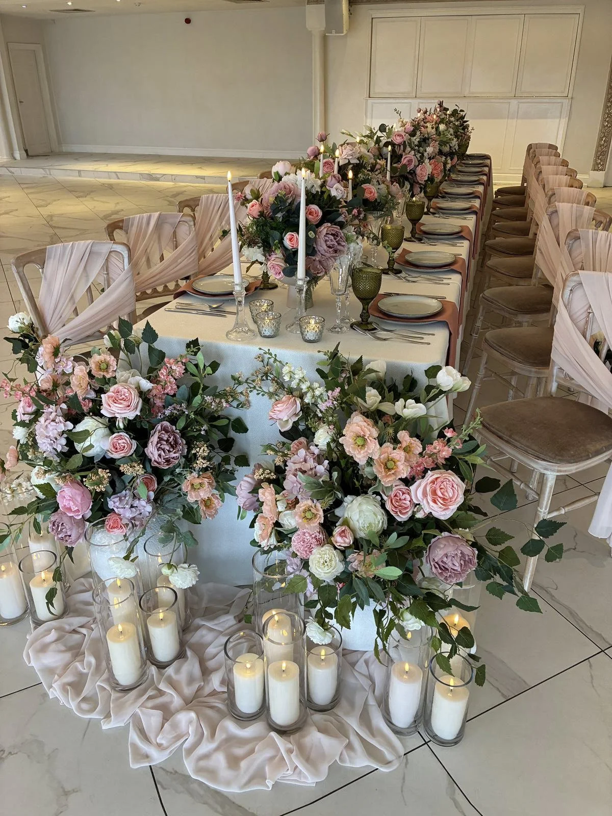 Cylinder Vase - 40cm Wide holding white pillar candles, surrounded by floral arrangements and soft pink fabric, available to hire for your wedding or event.