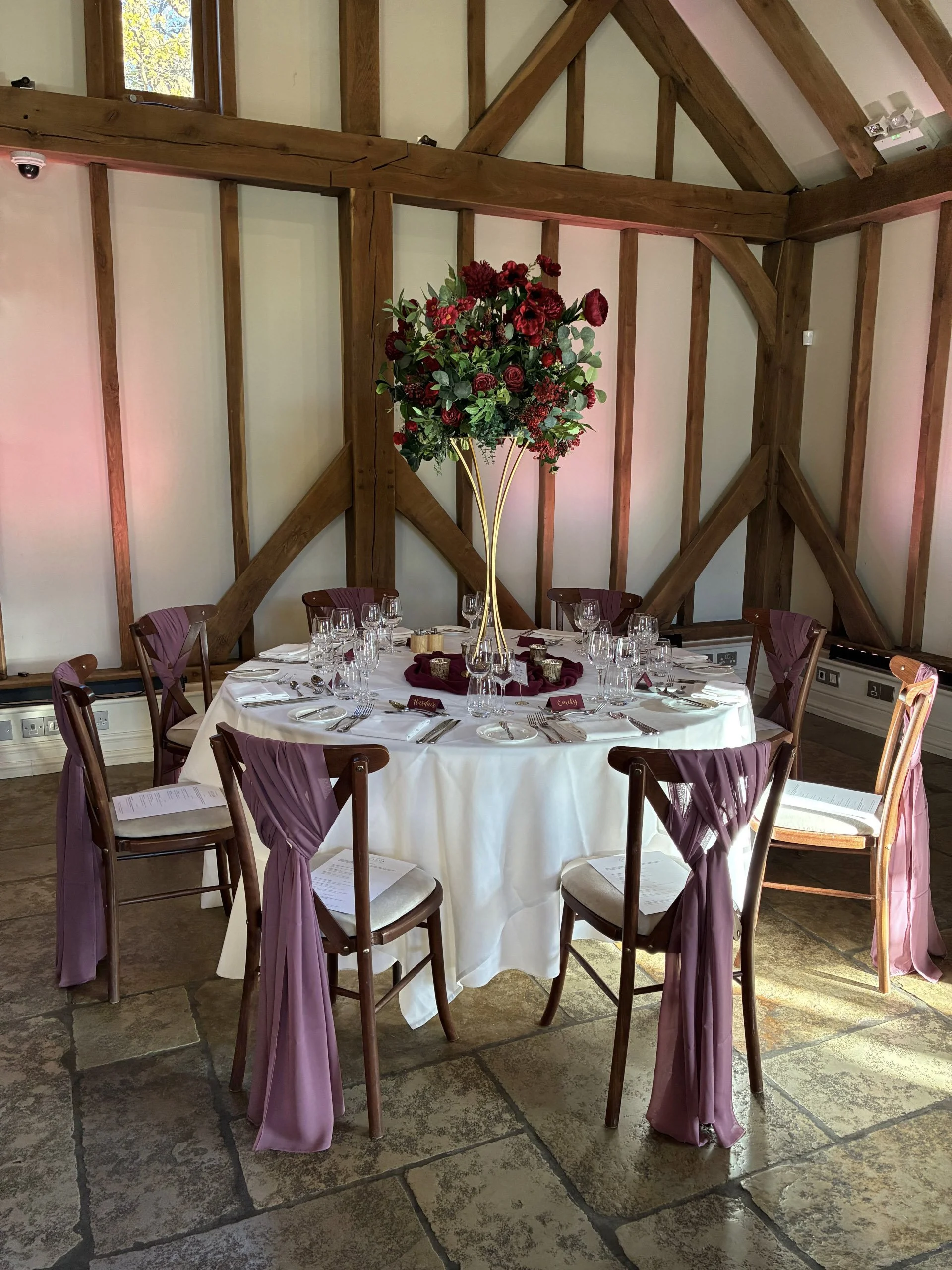 Elegant Gold Floral Stand - 76cm high 20cm top with deep red floral arrangement, available to hire for your wedding or event, displayed as a table centerpiece in a rustic venue.