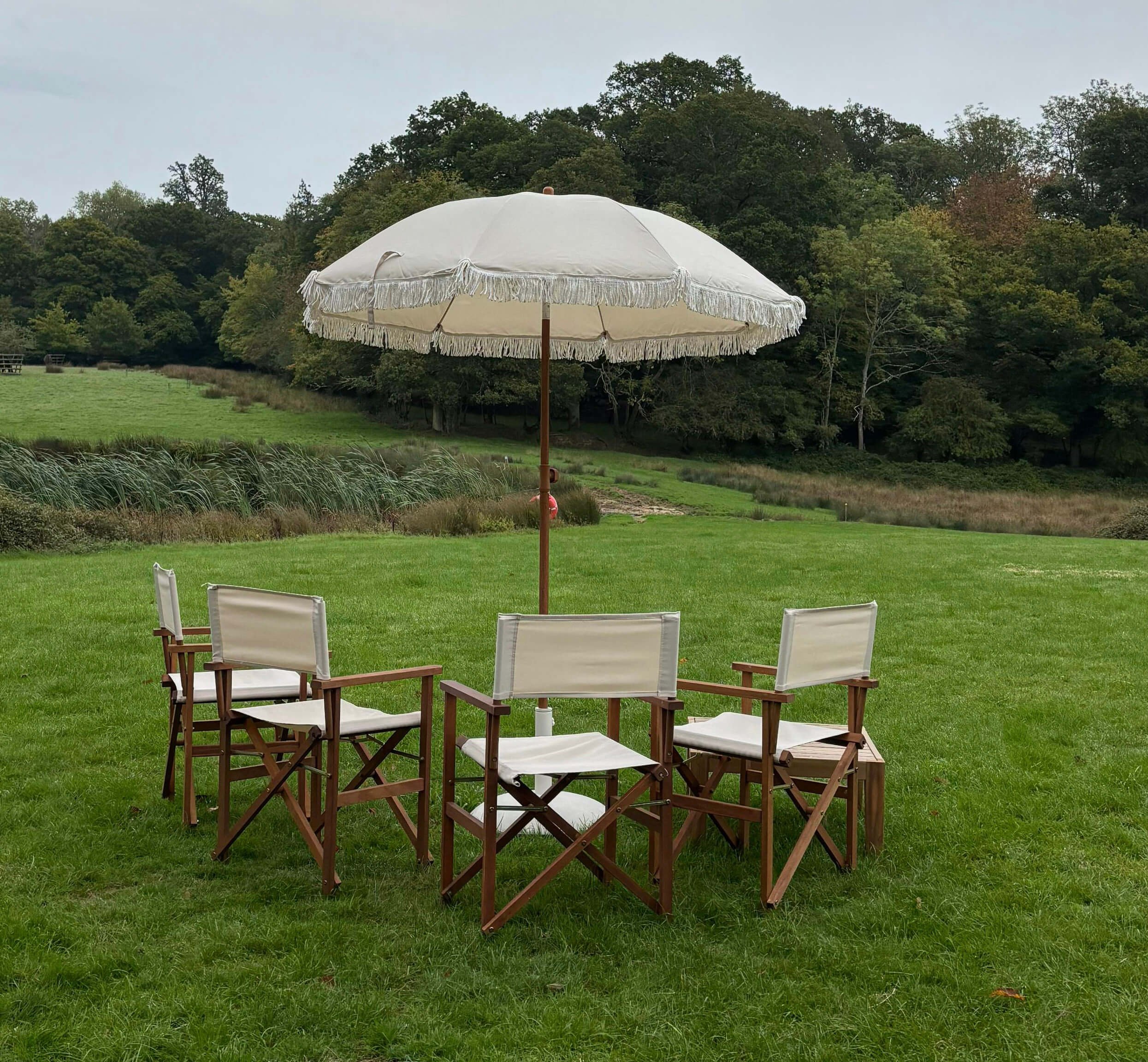 Directors Chair - Cream with wooden frame arranged outdoors under a cream fringed parasol, available to hire for your wedding or event.