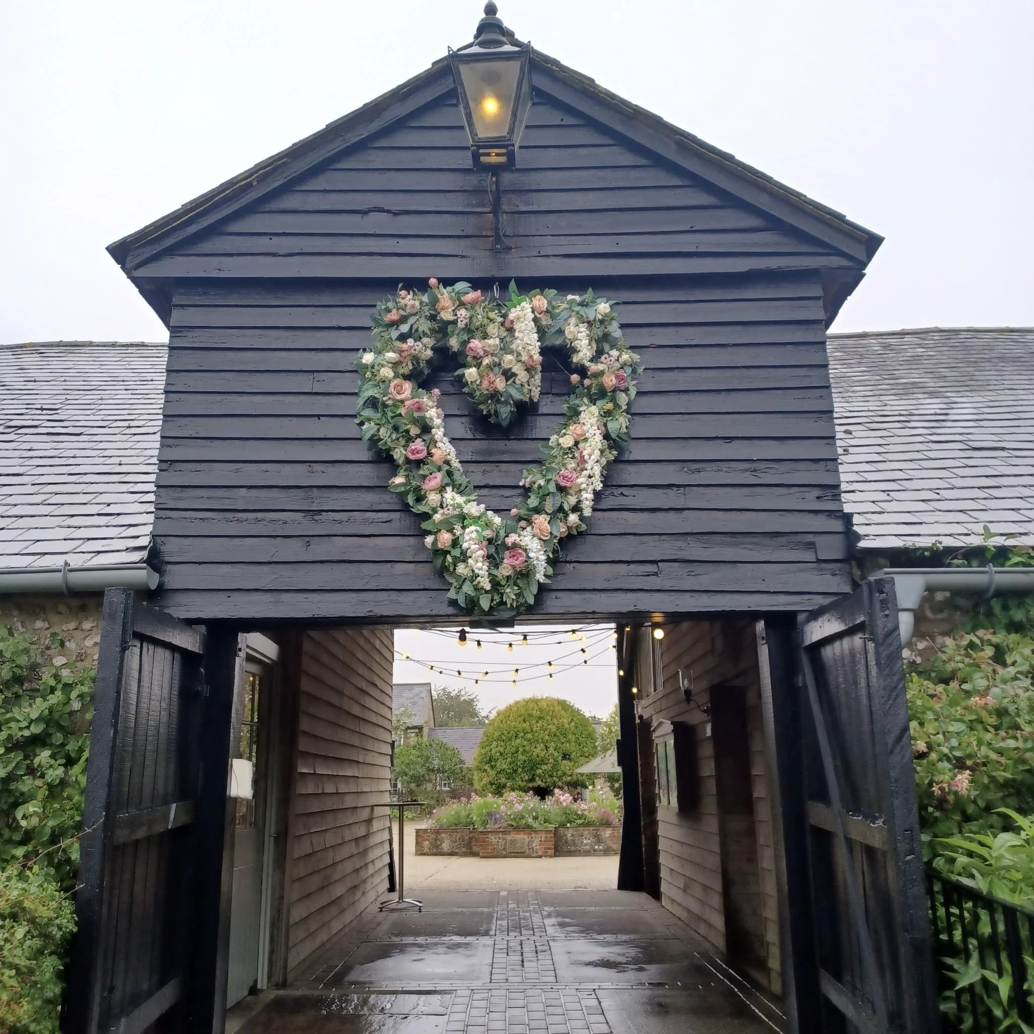 Giant Heart floral wreath in pink, white, and cream with greenery, available to hire for your wedding or event, displayed on a dark wooden building.