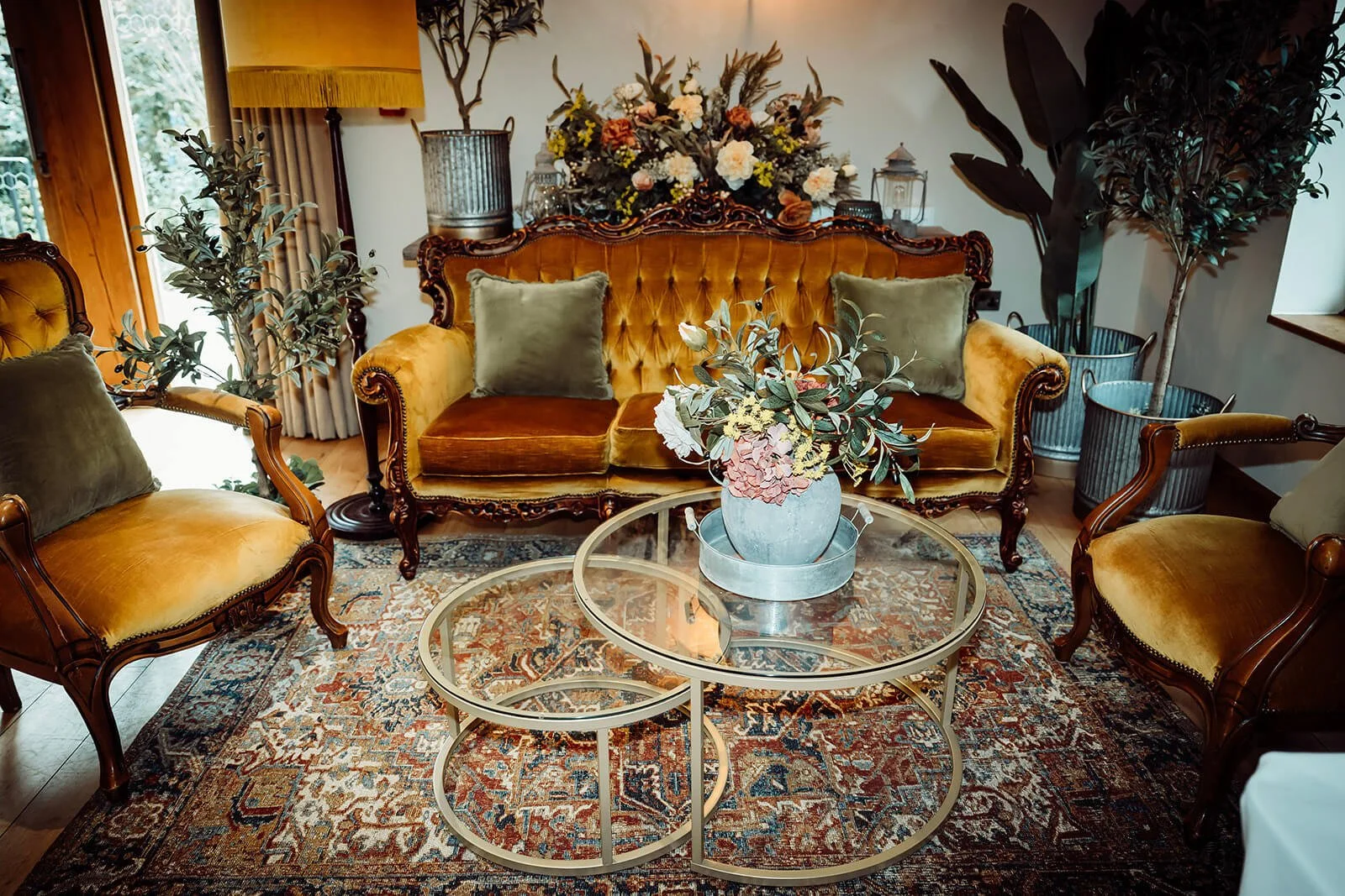 Gold Glass Coffee Table set of 2 with floral arrangement on top, nested on a patterned rug in a vintage-style room, available to hire for your wedding or event.