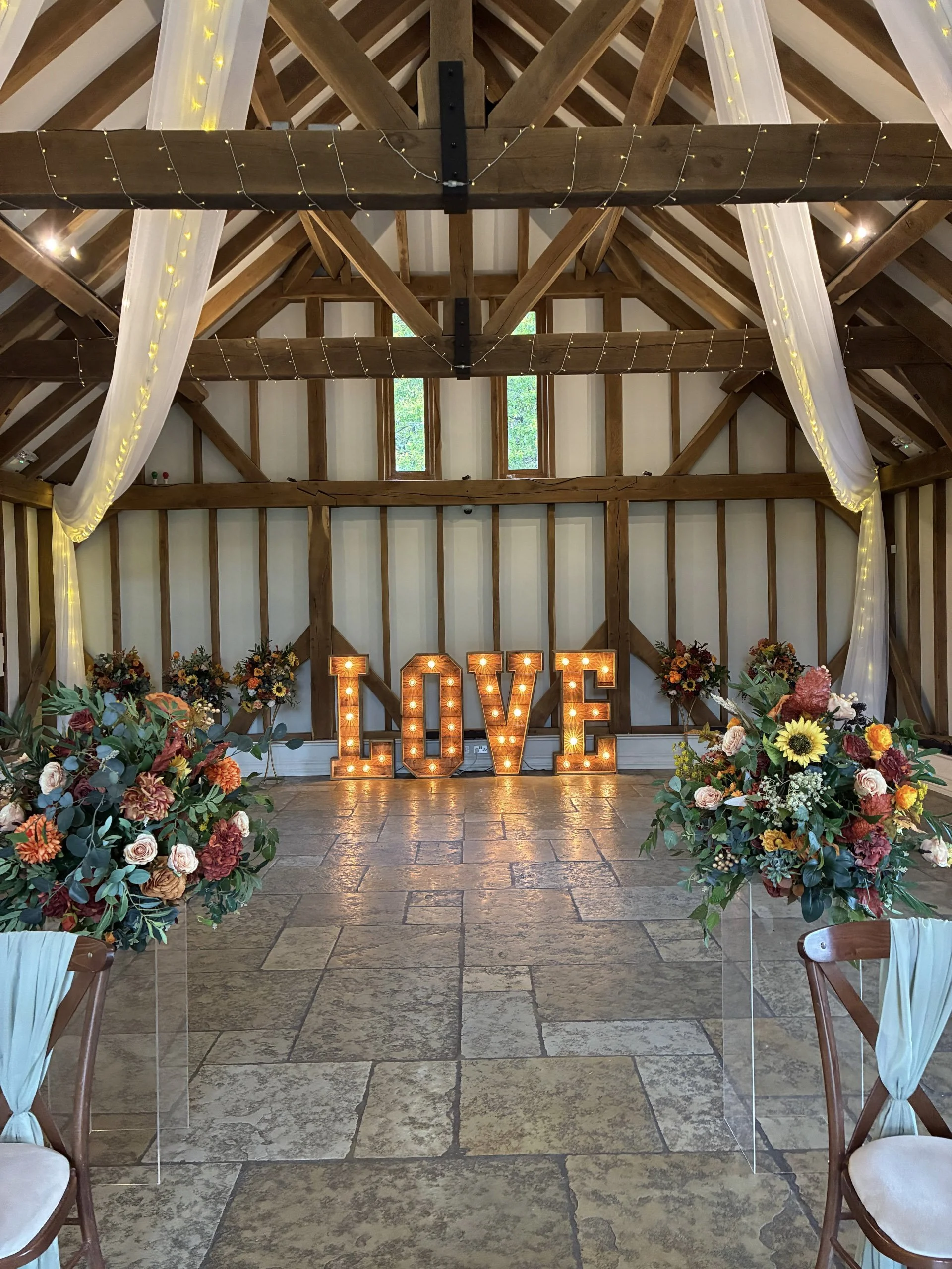 Giant Light Up Letters Rustic Love with warm bulbs in a barn setting, surrounded by floral arrangements and draped fabric, available to hire for your wedding or event.