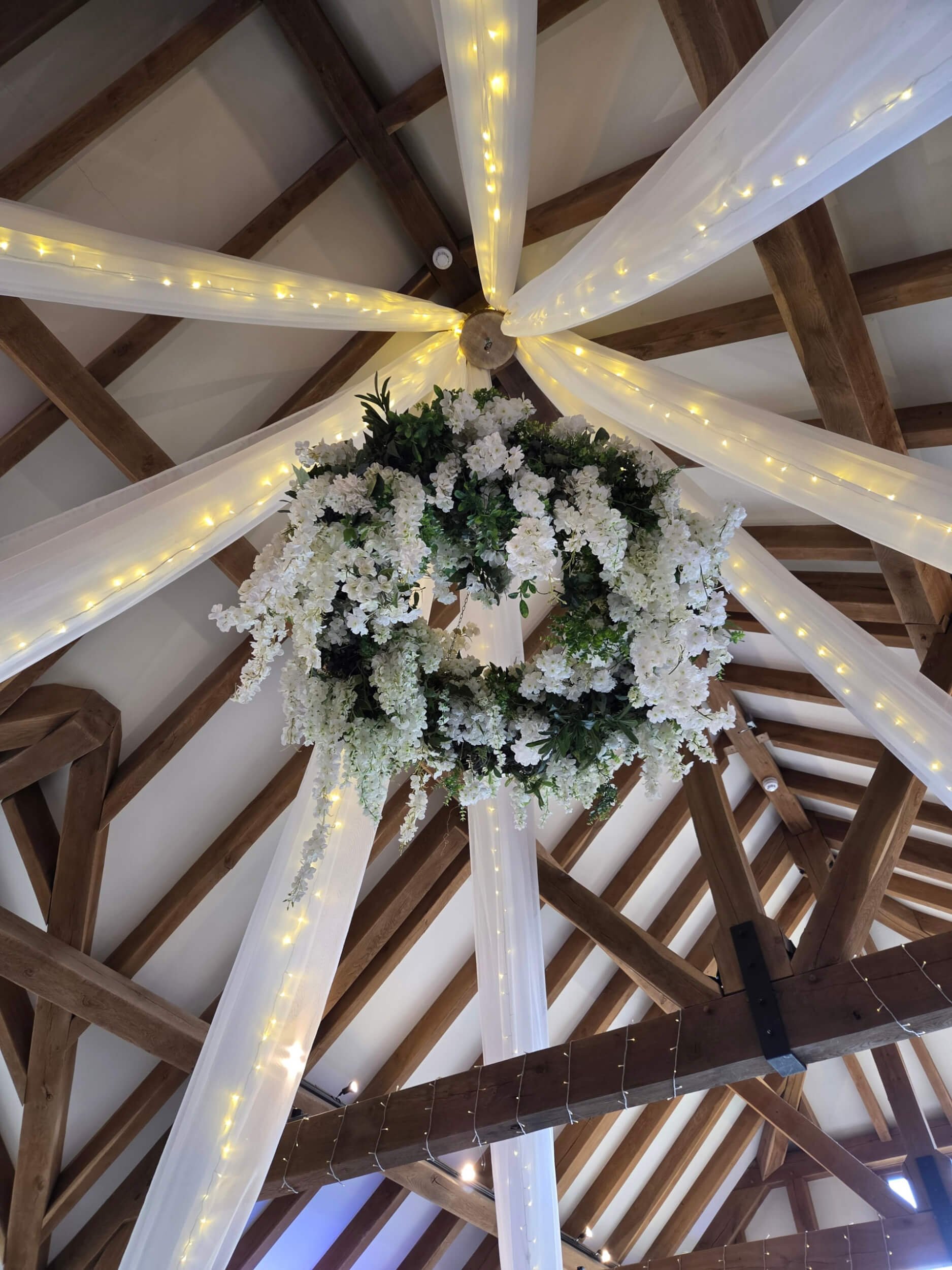 Wisteria Chandelier with white and green flowers and fairy lights, draped with white fabric, available to hire for your wedding or event.