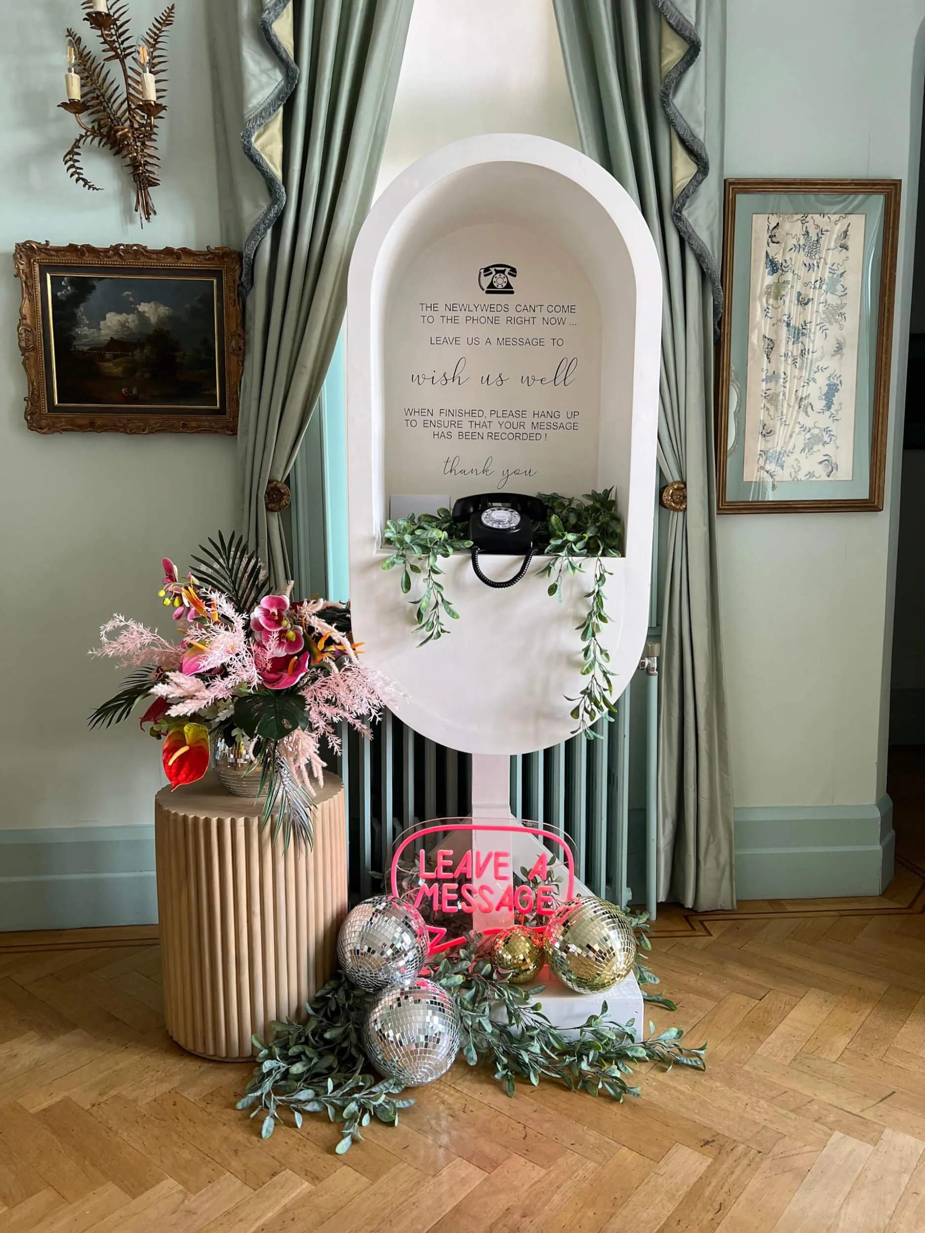 Audio Phone Booth with vintage rotary phone and greenery, decorated with disco balls and neon 'Leave a Message' sign, available to hire for your wedding or event.