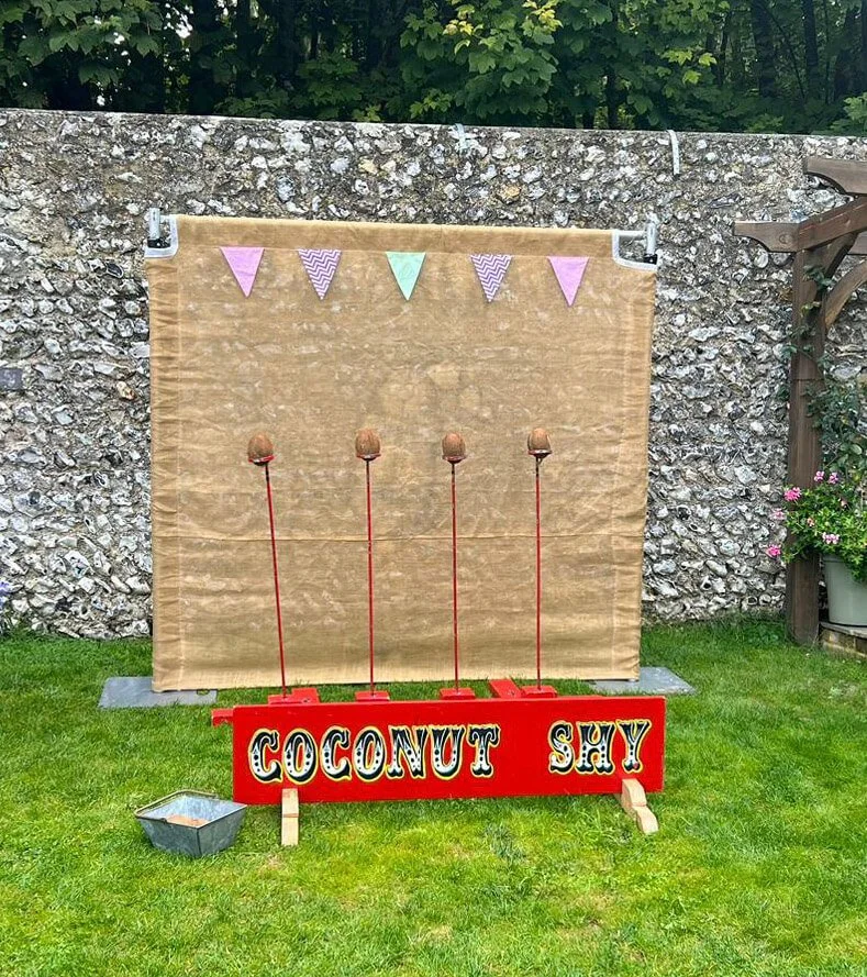 Coconut Shy game with four coconuts on red poles and a beige backdrop with bunting, available to hire for your wedding or event.