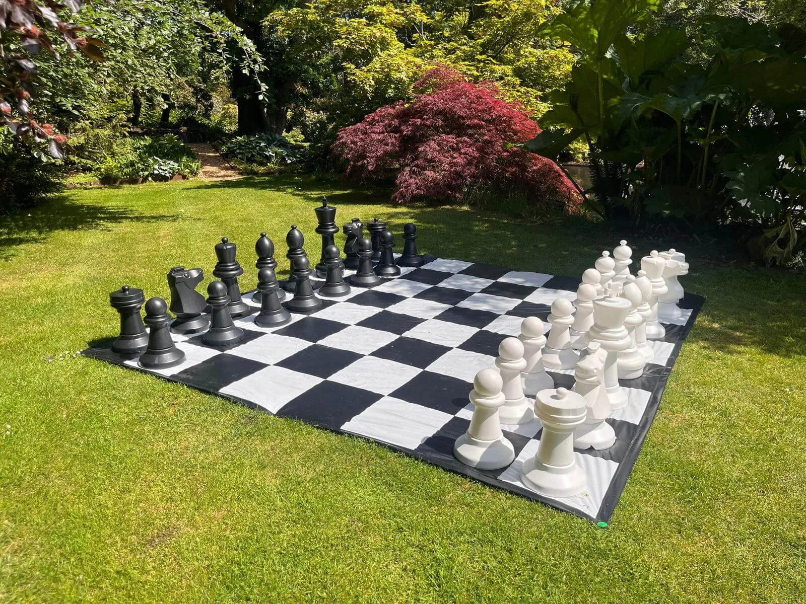 Giant Chess set with large black and white pieces on a checkered mat, available to hire for your wedding or event in a garden setting.