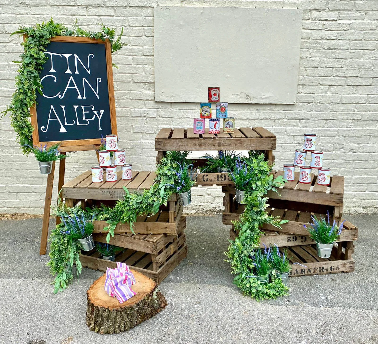Tin Can Alley game with numbered tin cans stacked on wooden crates decorated with greenery, available to hire for your wedding or event.