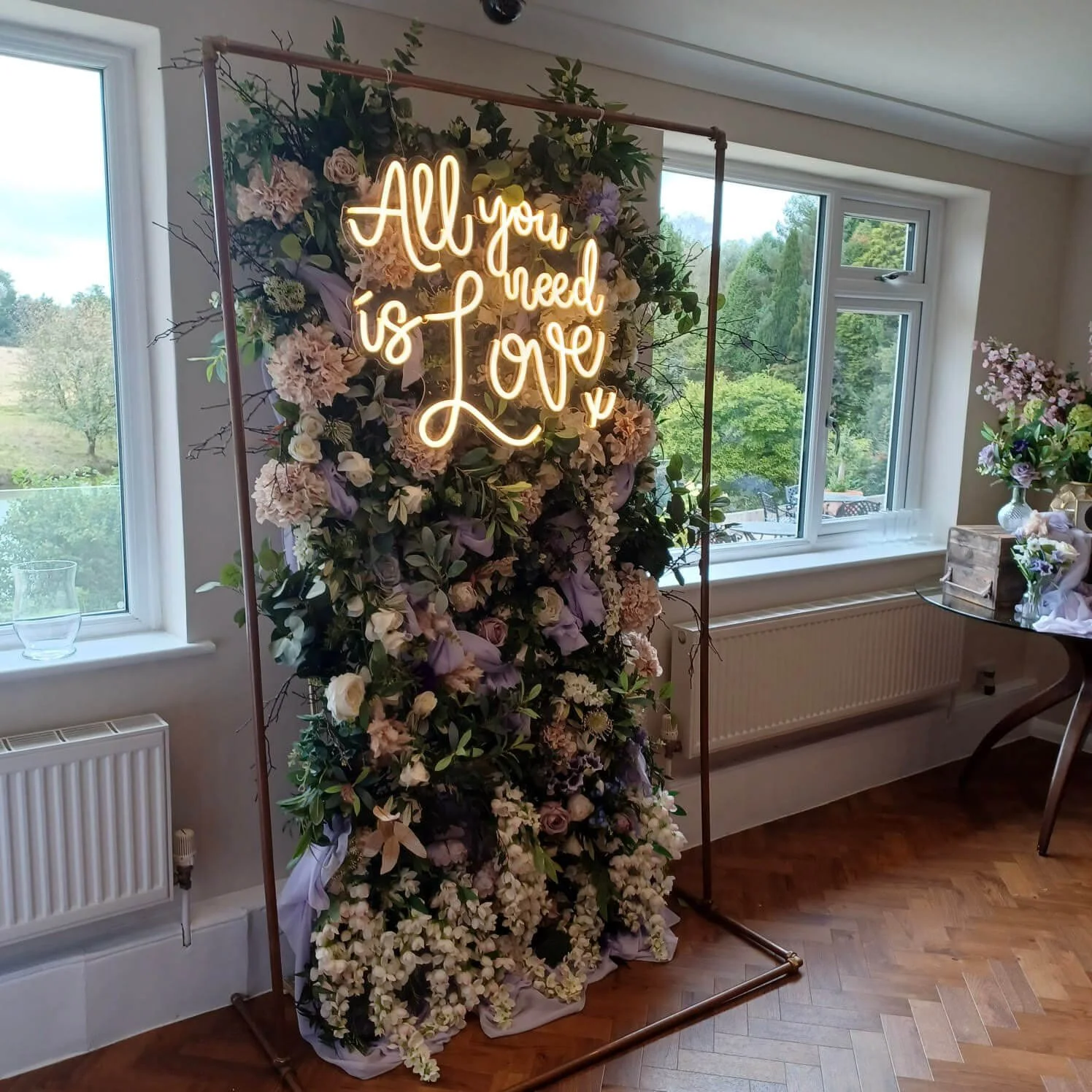 All You Need is Love Neon Sign illuminated on a floral backdrop, available to hire for your wedding or event.