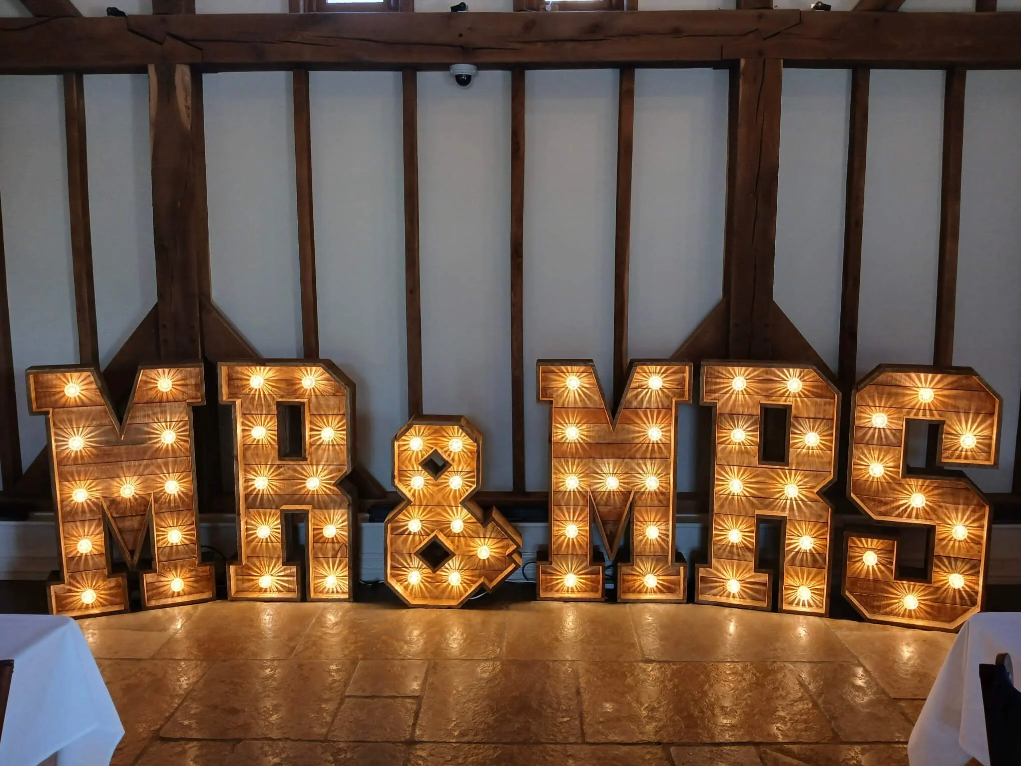 Giant Light Up Letters - Mr & Mrs - RUSTIC with warm bulbs, available to hire for your wedding or event, displayed indoors against a white wall with wooden beams.