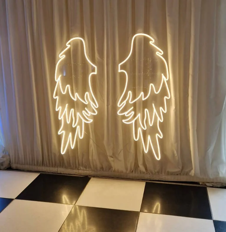 Angel Wings Neon Sign glowing white against a cream curtain backdrop, available to hire for your wedding or event.