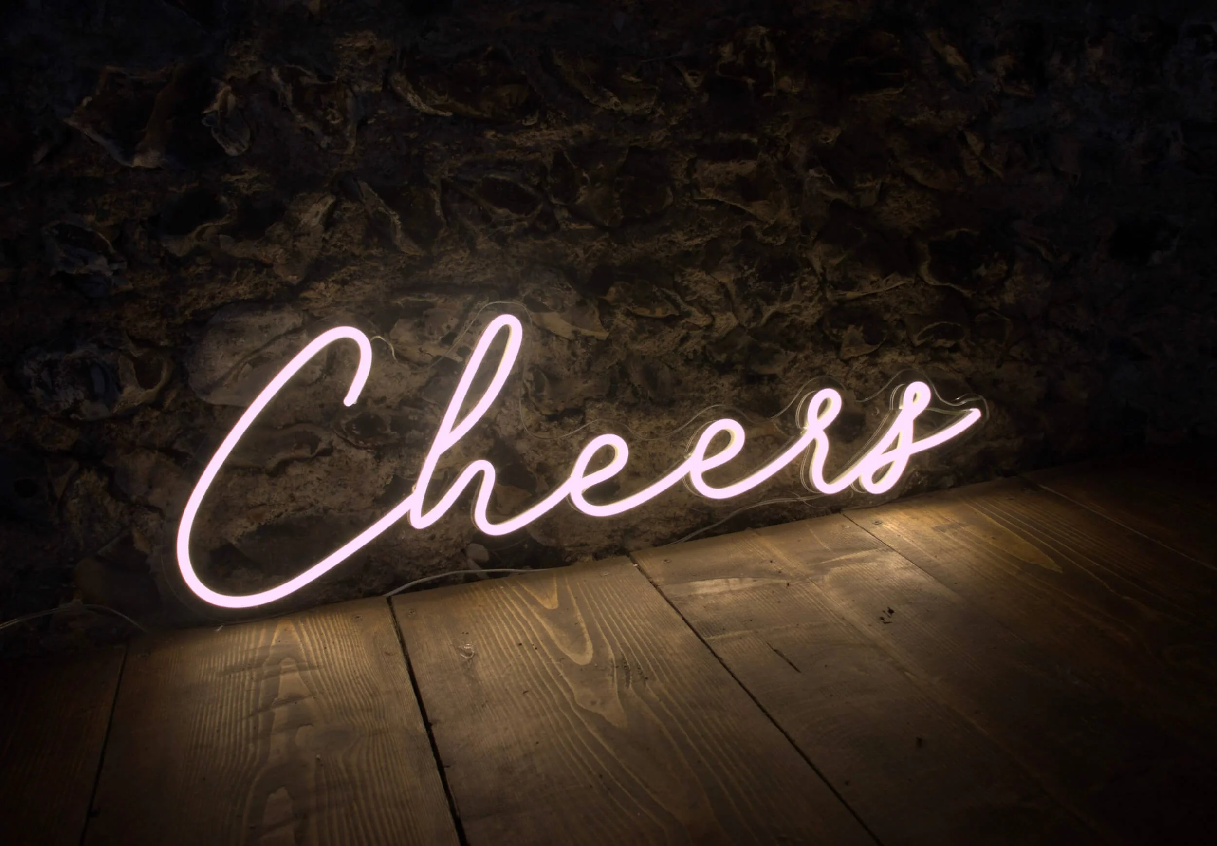 Cheers Neon Sign in pink neon light, placed on wooden floor against dark stone wall, available to hire for your wedding or event.