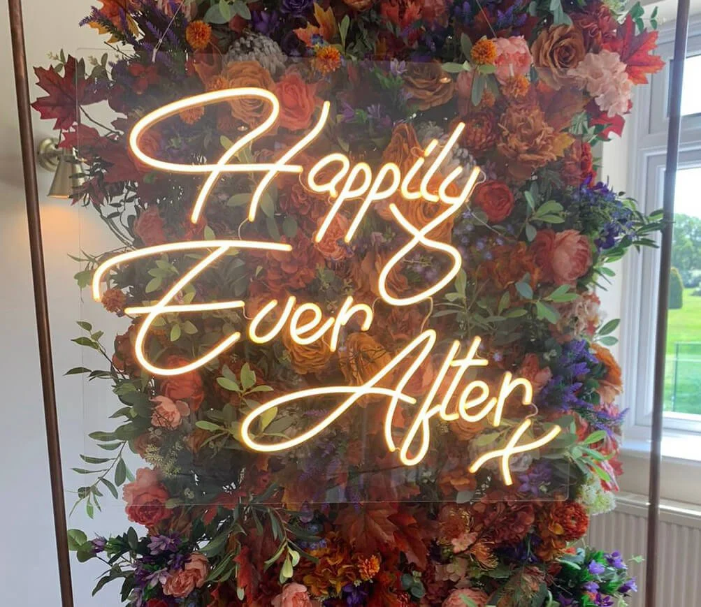 Happily Ever After Neon Sign in warm white, mounted on clear panel with floral backdrop, available to hire for your wedding or event.