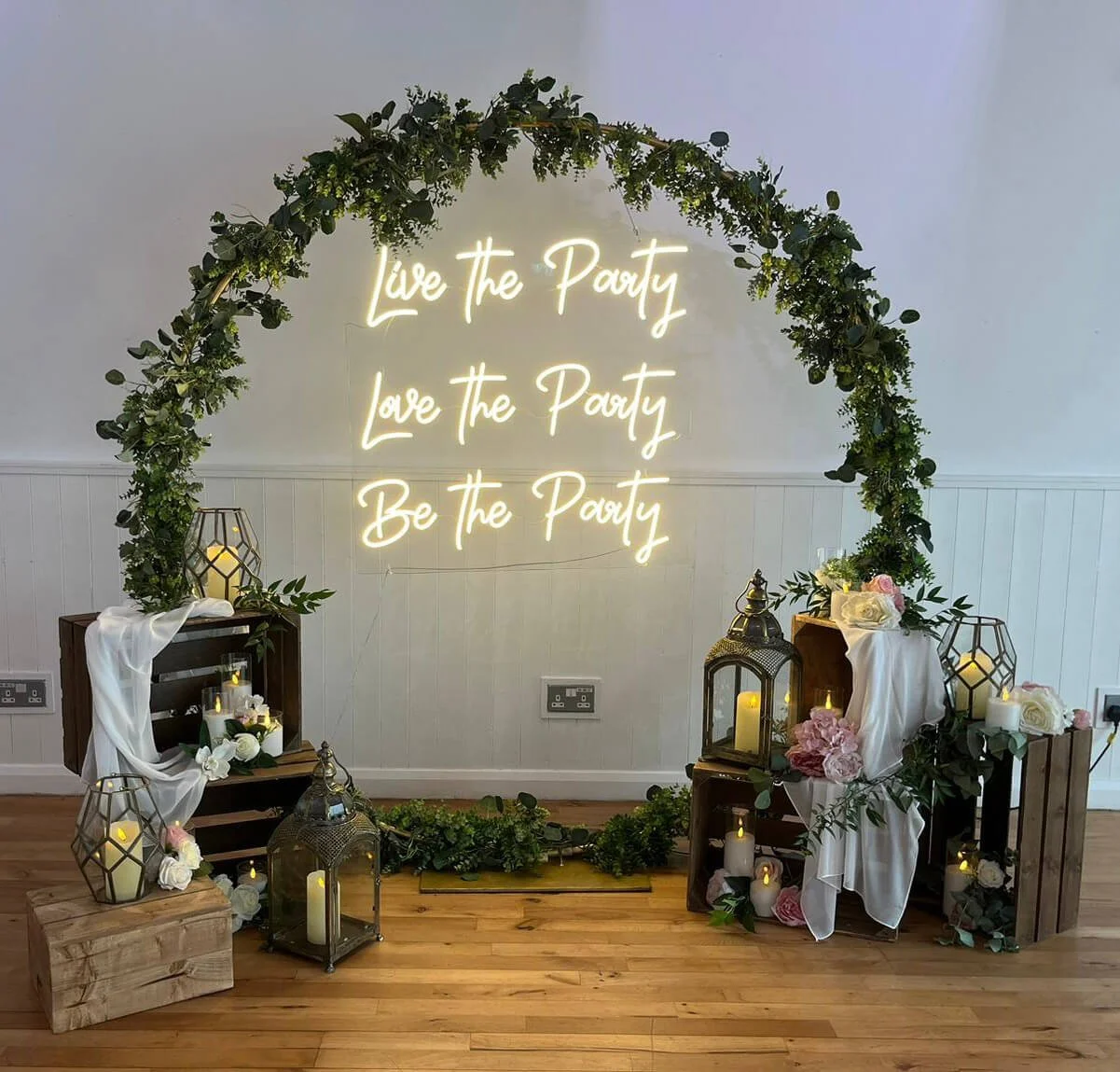 Live the Party, Love the Party, Be the Party Neon Sign glowing white on a foliage-covered arch with lanterns and candles, available to hire for your wedding or event.