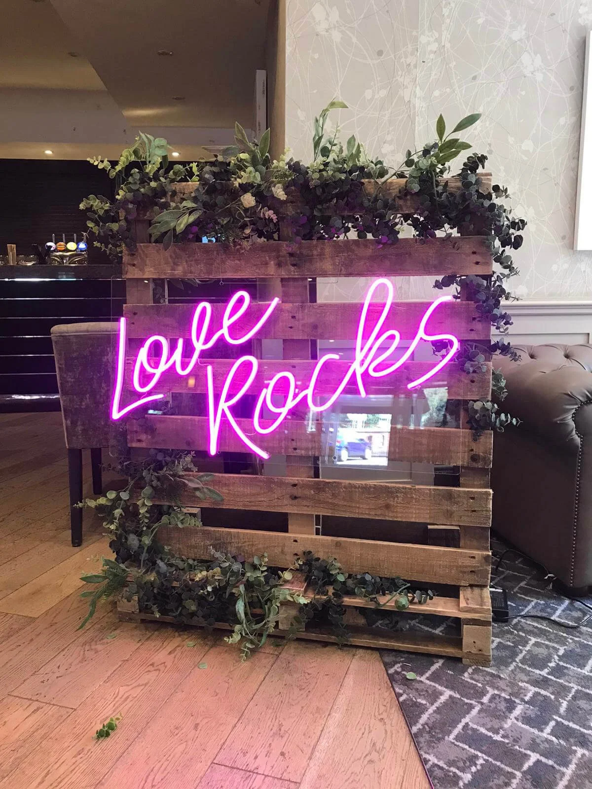 Love Rocks Neon Sign in bright pink neon on a wooden pallet with greenery, available to hire for your wedding or event.