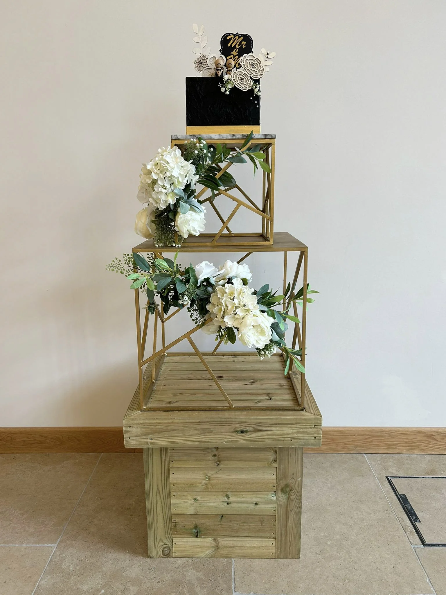 Geo Gold Cake Stand - 30cm square with white floral decorations and a black cake on top, available to hire for your wedding or event.