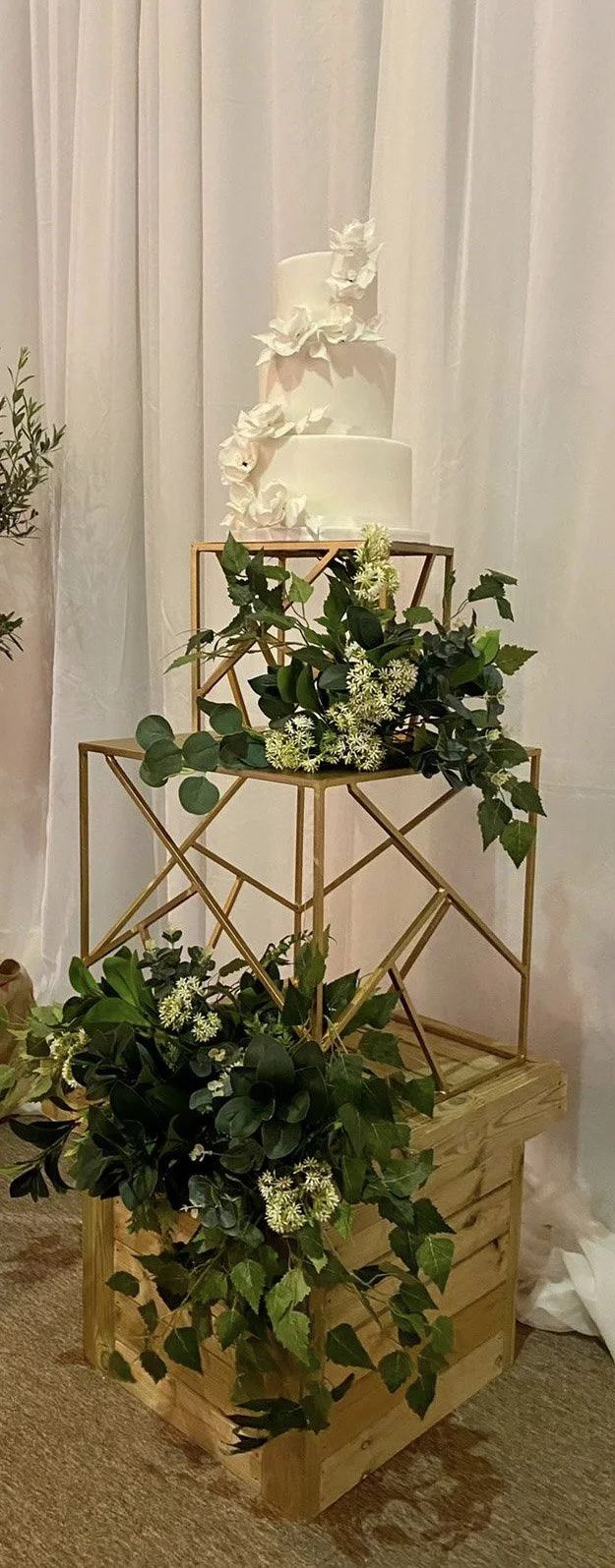 Geo Gold Cake Stand - 50cm square holding a three-tier white wedding cake with floral decoration, available to hire for your wedding or event.