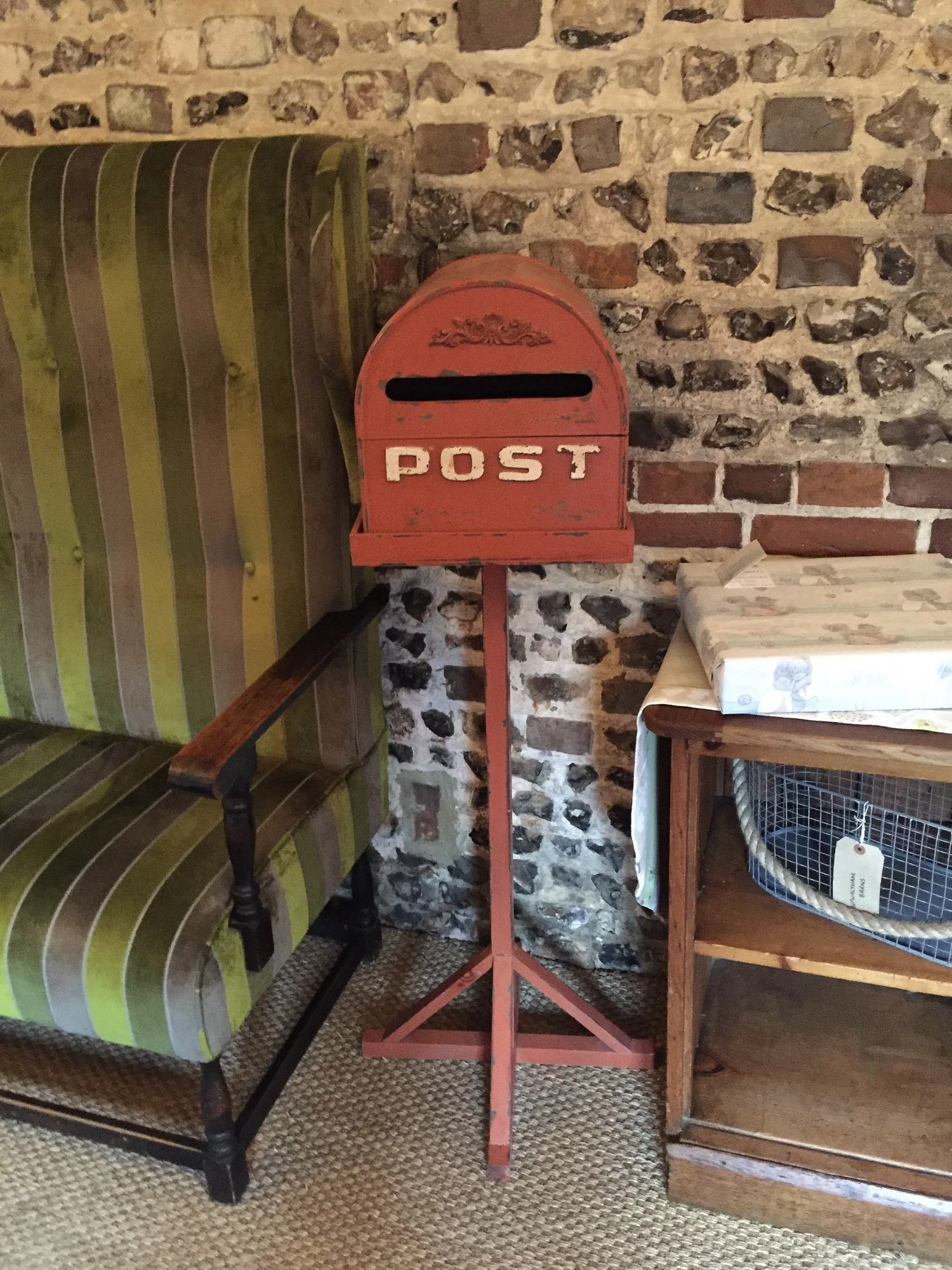 Rustic Red Post Box in red with white lettering on a stand, available to hire for your wedding or event, shown indoors by a stone wall and vintage furniture.