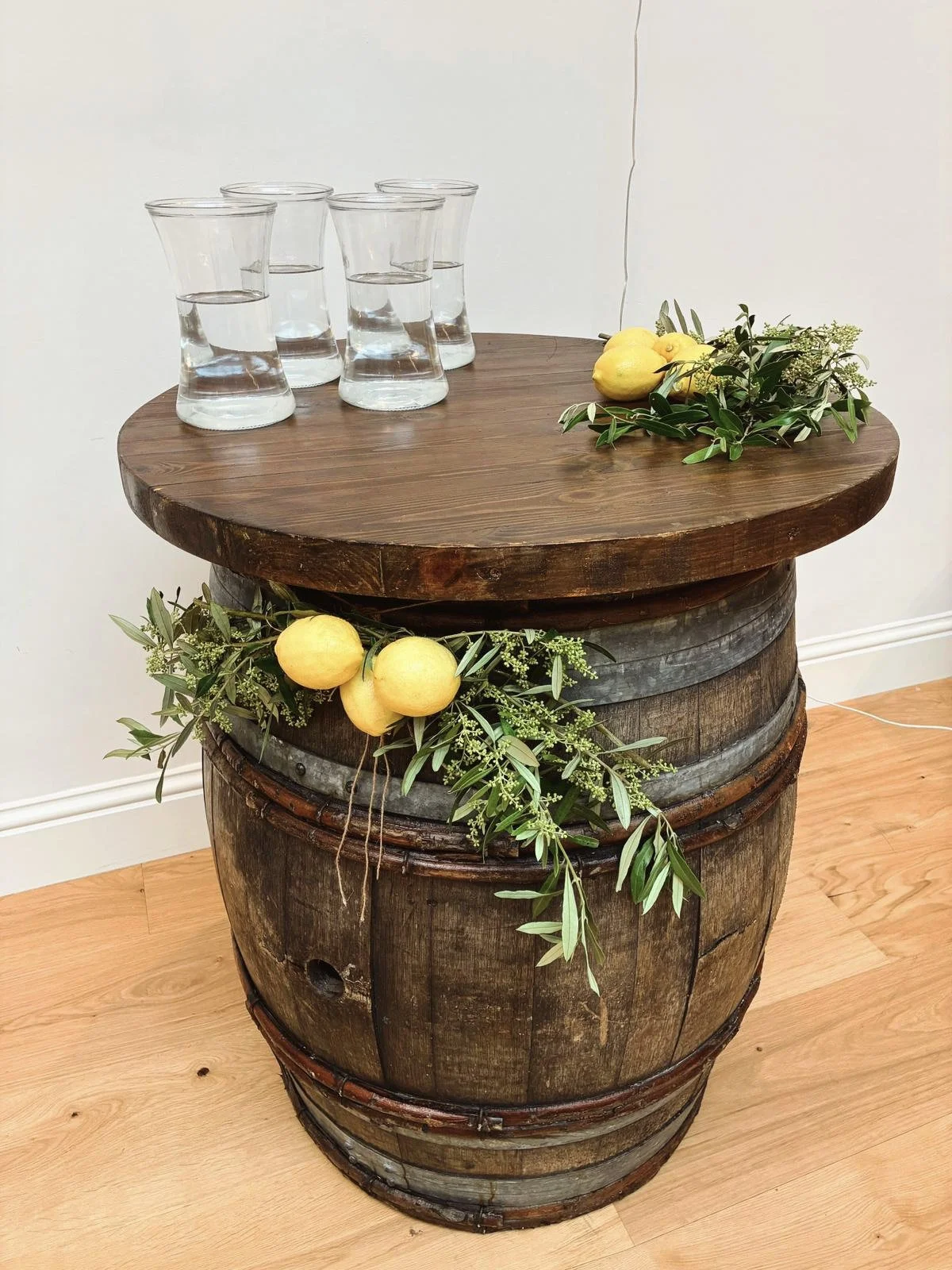 Barrel with oak top decorated with lemons and greenery, holding four glasses of water, available to hire for your wedding or event.