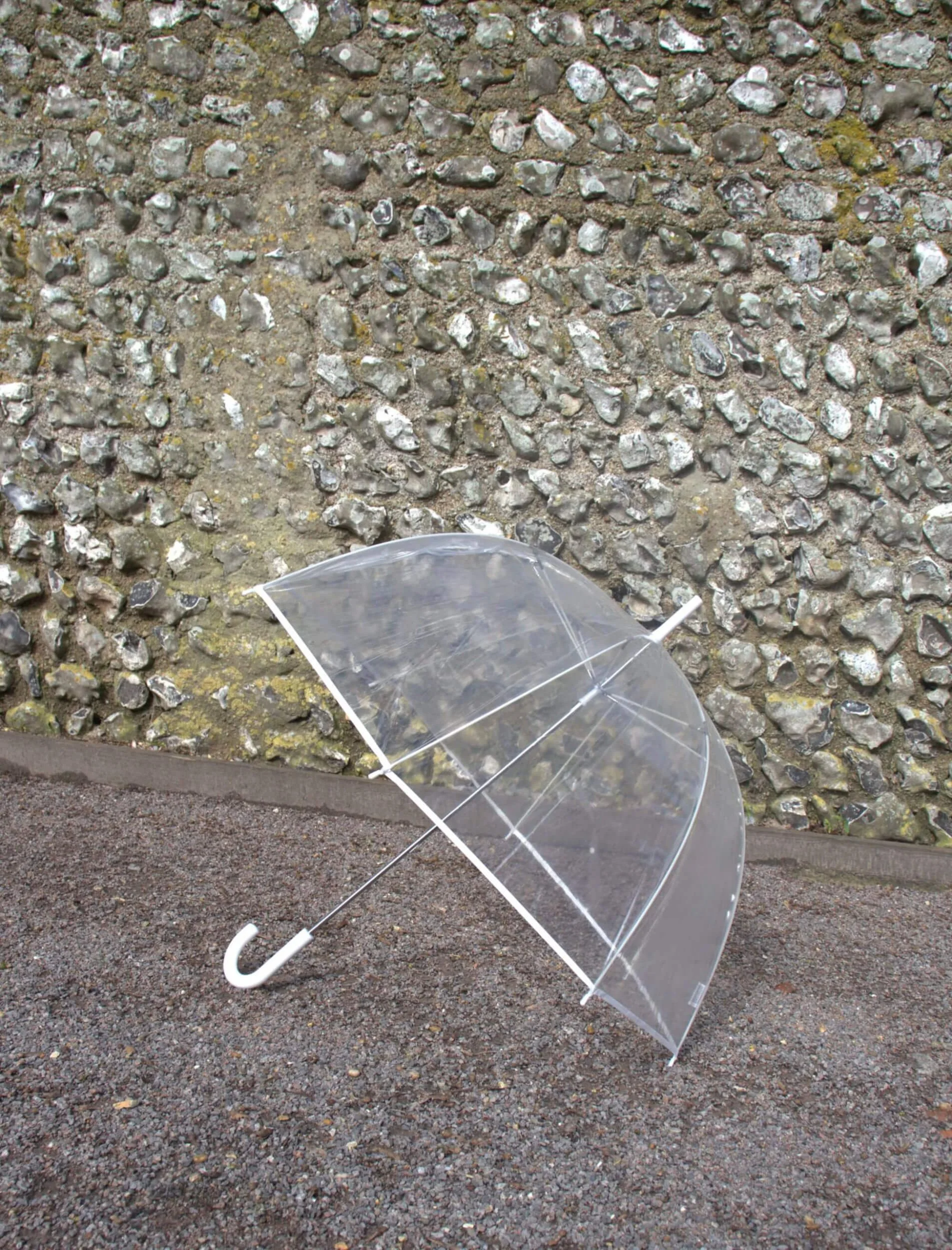 Clear Dome Umbrellas with white handle and ribs, available to hire for your wedding or event, shown open on gravel against a stone wall.